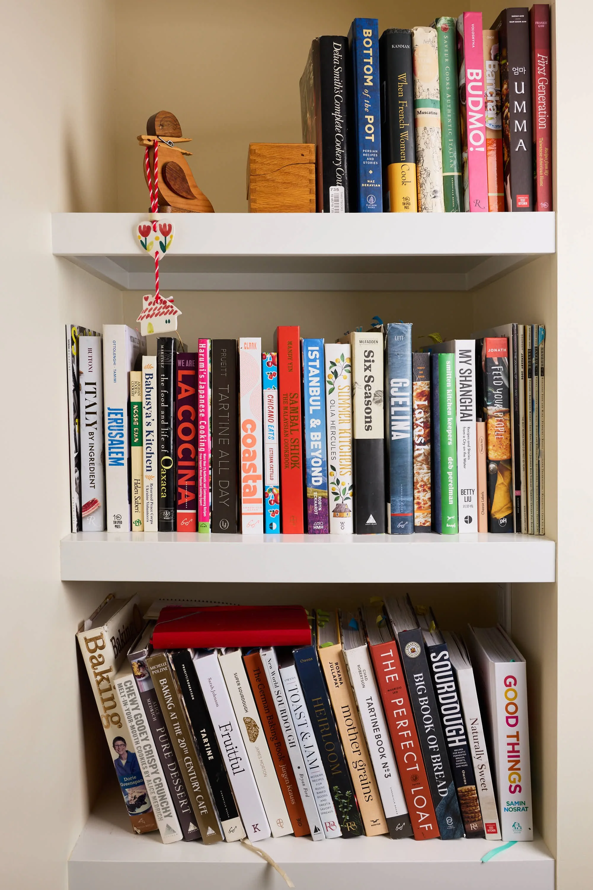 A white bookshelf with three shelves filled with a variety of colorful books. The top shelf has books with titles related to cooking and food, and features a small wooden bird figurine with a red and white string hanging with decorative hearts and a house design. The middle shelf contains books on travel, culture, and cuisine, with some horizontal books visible at the bottom. The bottom shelf contains an assortment of books on food, history, and other topics, with some books leaning to the side and a piece of string or ribbon hanging from the right side.