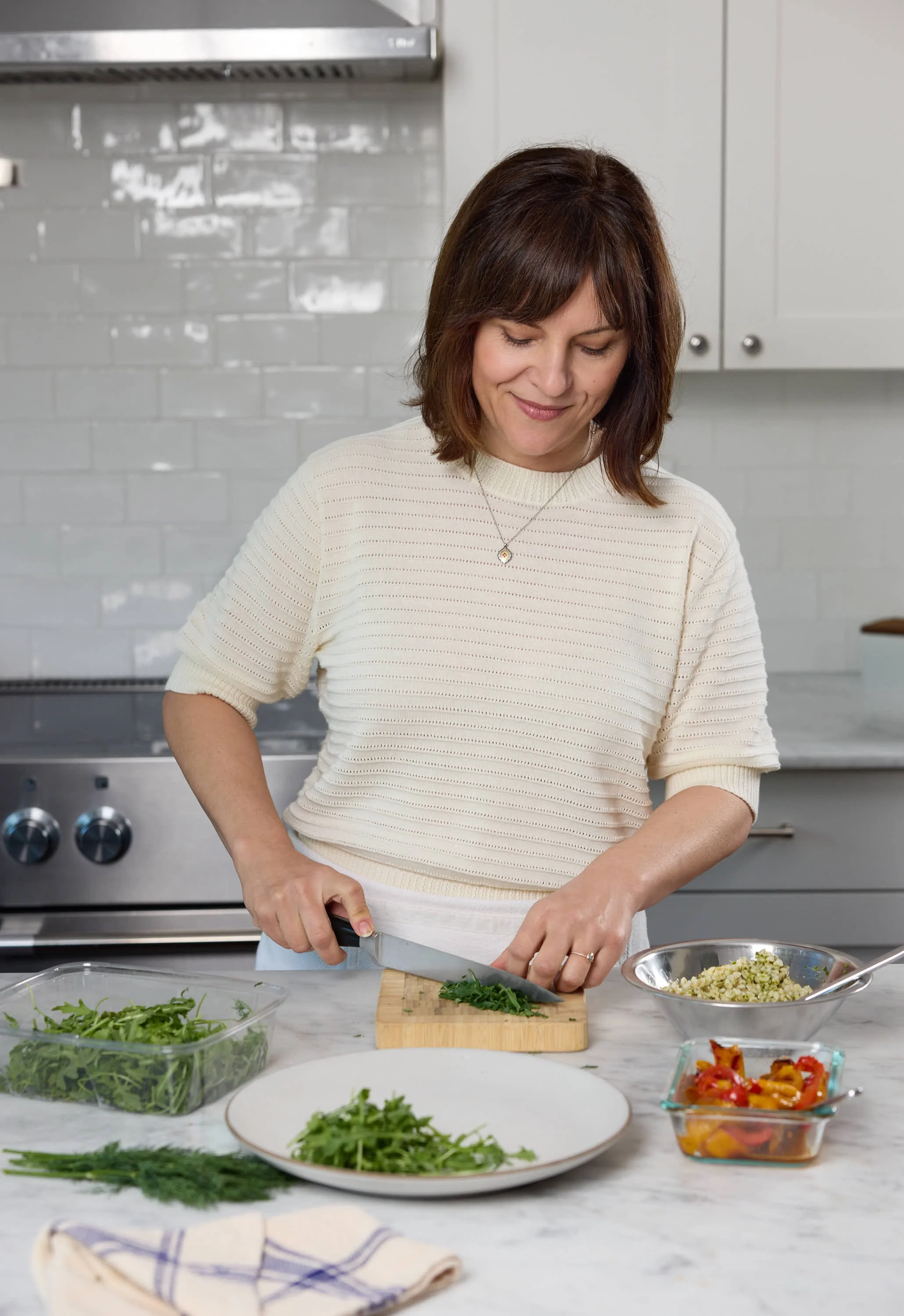 Woman chopping greens on a wooden cutting board in a modern kitchen surrounded by fresh vegetables and herbs.
