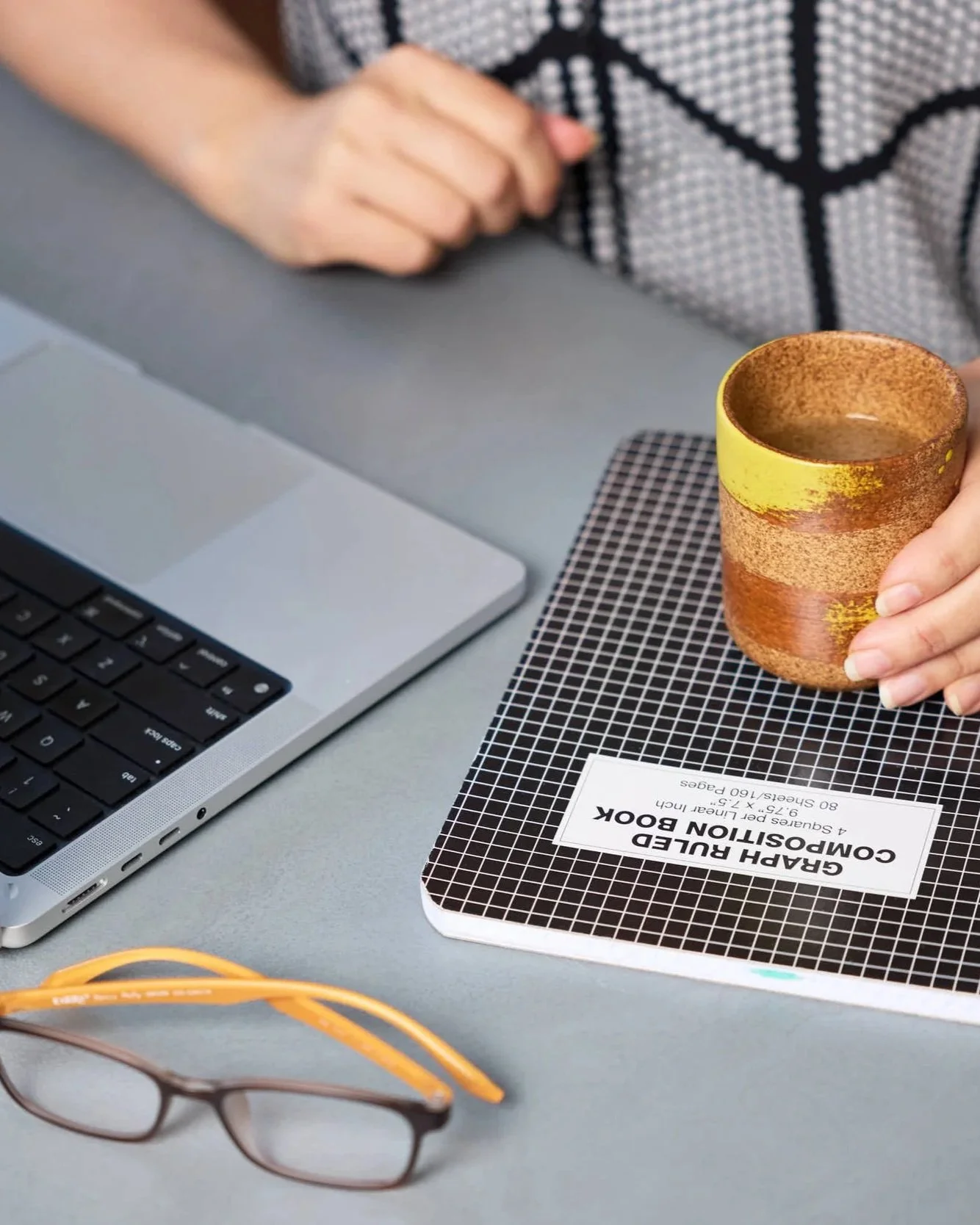 A person holding a yellow and brown ceramic cup on a desk with a laptop, a black and white grid notebook labeled 'GRAH RULER COMPASSION BOOK,' a pair of glasses, and yellow glasses cords.