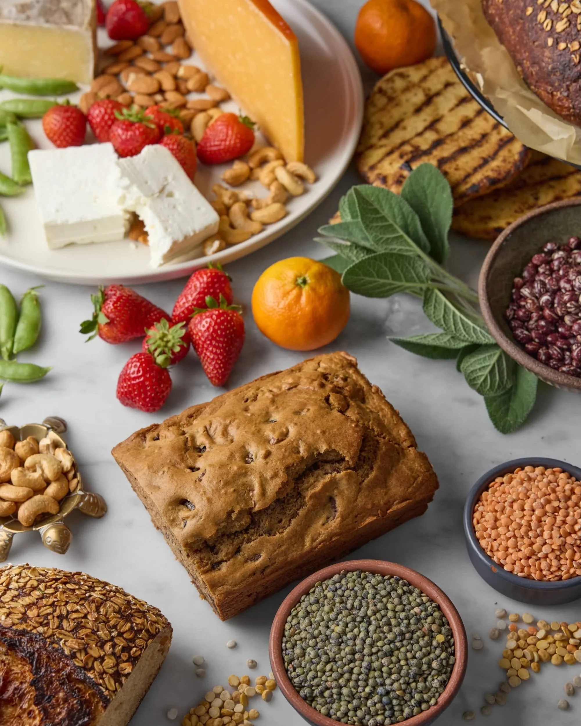 Assorted baked goods, cheeses, strawberries, and various grains and seeds displayed on a surface.
