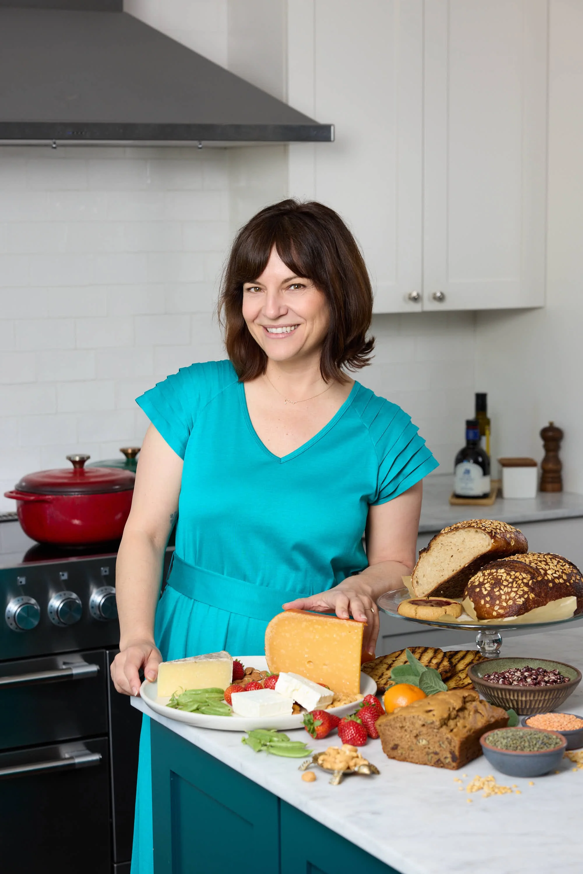 Woman in blue dress standing behind a kitchen counter with bread, cheese, fruit, and baked goods, smiling at camera.