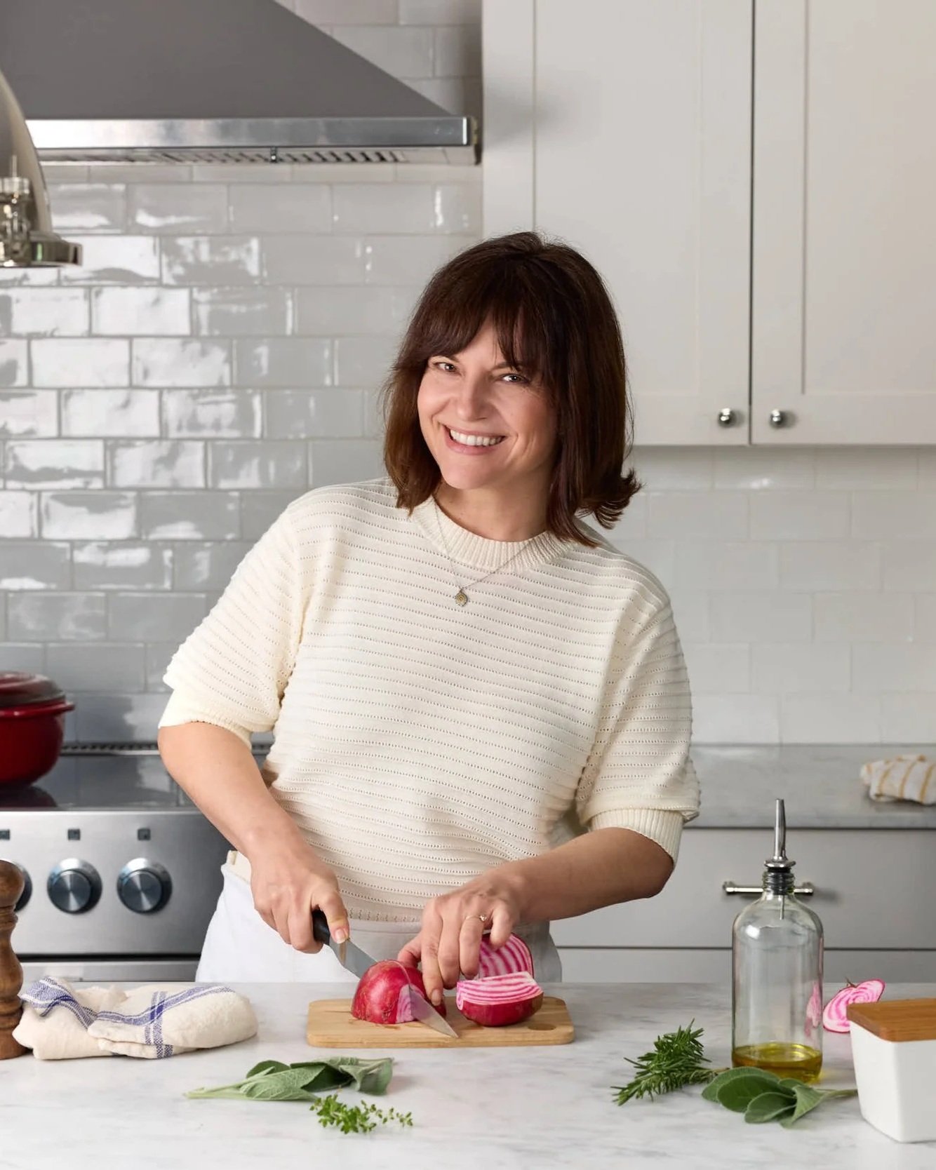 A woman with brown hair and a white sweater chopping an onion in a kitchen. There are fresh herbs, a bottle of olive oil, and a bowl with a lid on a white countertop.