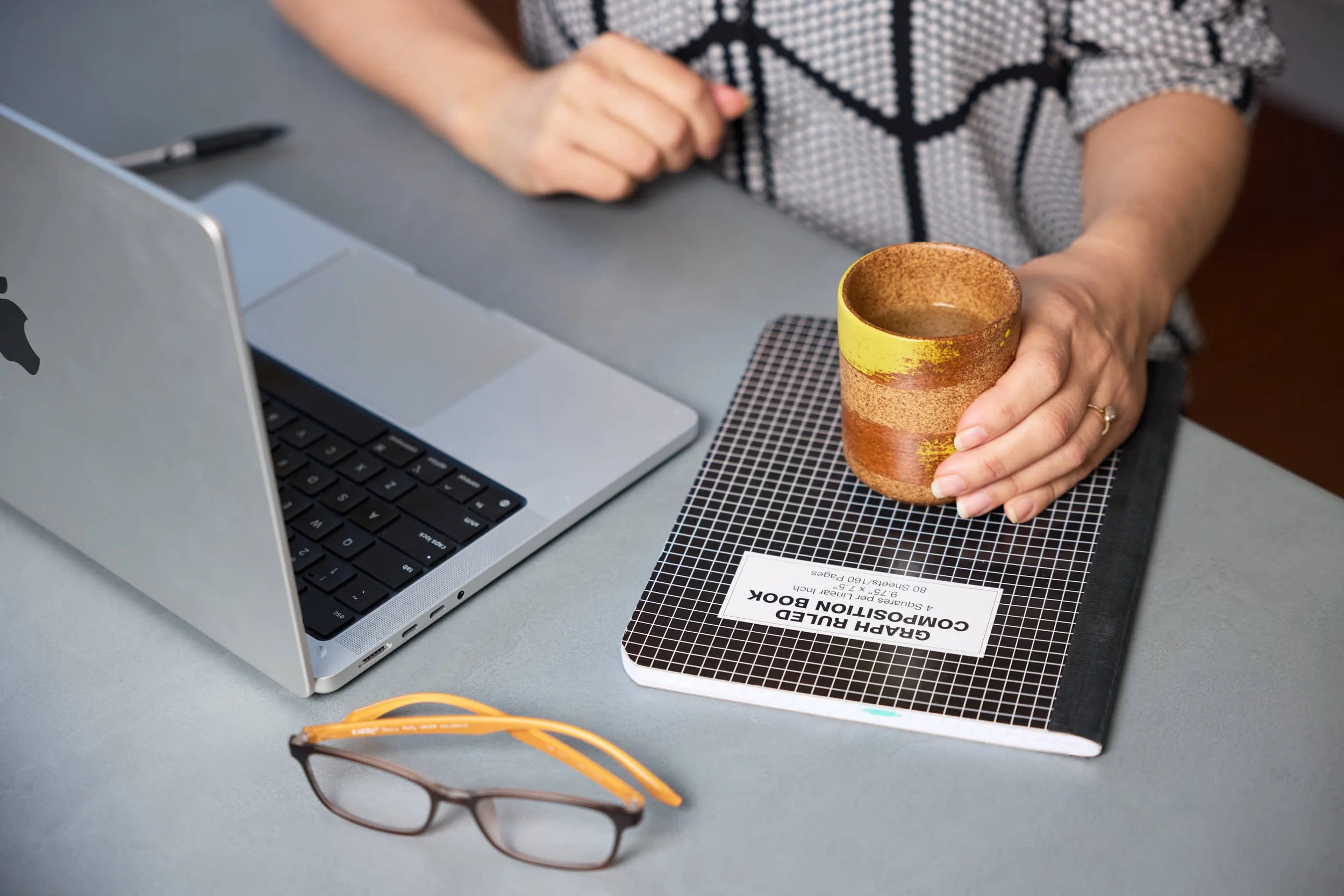 A person sitting at a desk holding a ceramic mug with a textured, two-tone yellow and brown design. On the desk, there is a silver laptop, a black pen, a pair of glasses with black frames, and a black-and-white grid-patterned notebook labeled "GRAH RULER BOOK" for a compass projection. There is also a sheet of paper with text about a seminar or workshop. The person's right arm is visible, wearing a patterned shirt with geometric designs.