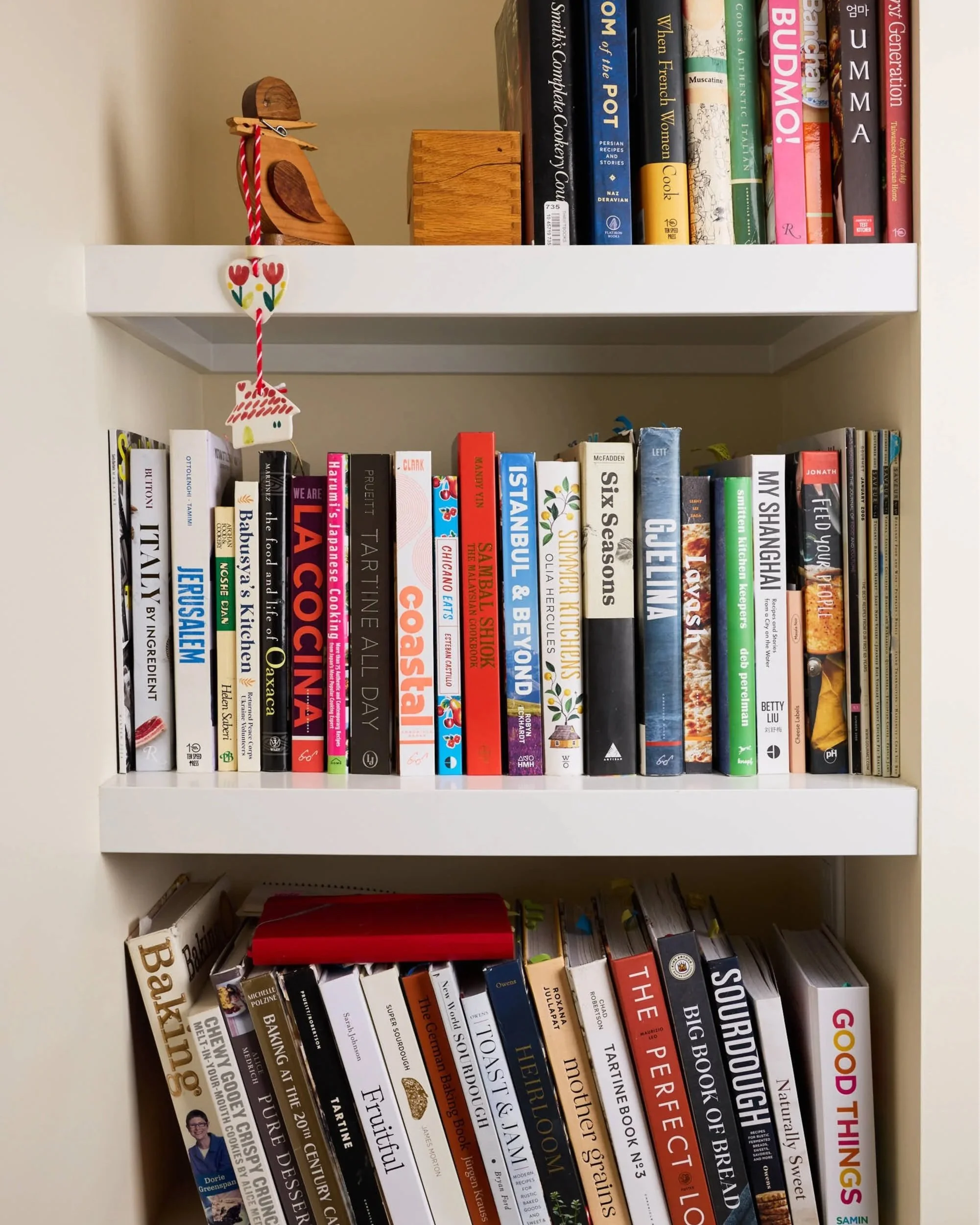 A white bookshelf with a row of cookbooks and interior design books, and decorative items such as a wooden bird figure, a small wooden box, and a hanging heart decoration with tulips. The books have colorful covers and are arranged side by side.