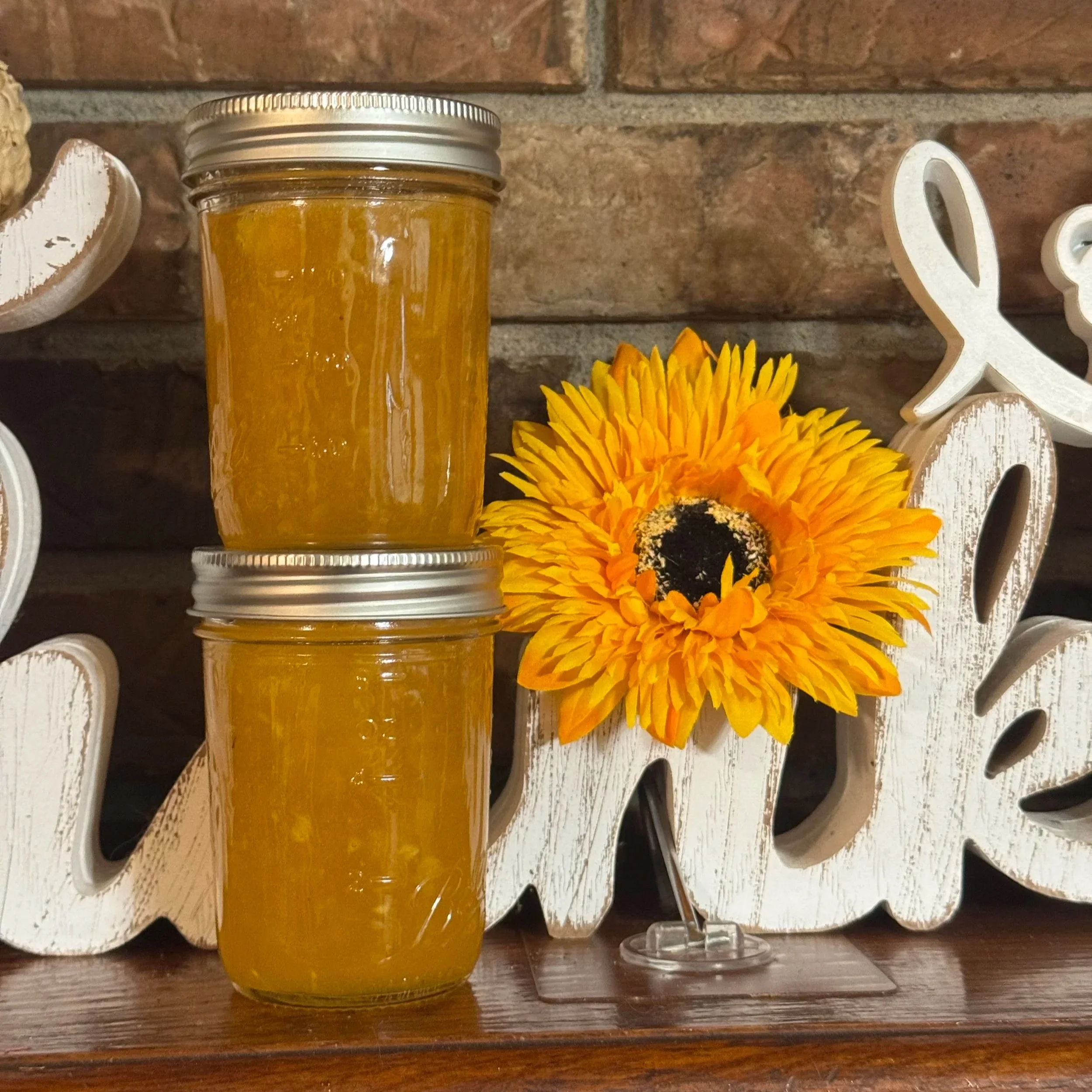 Two jars of honey stacked on top of each other, next to a large sunflower with yellow petals and dark center, on a wooden surface with a decorative white wooden sign that spells 'Love' in the background and a brick wall.