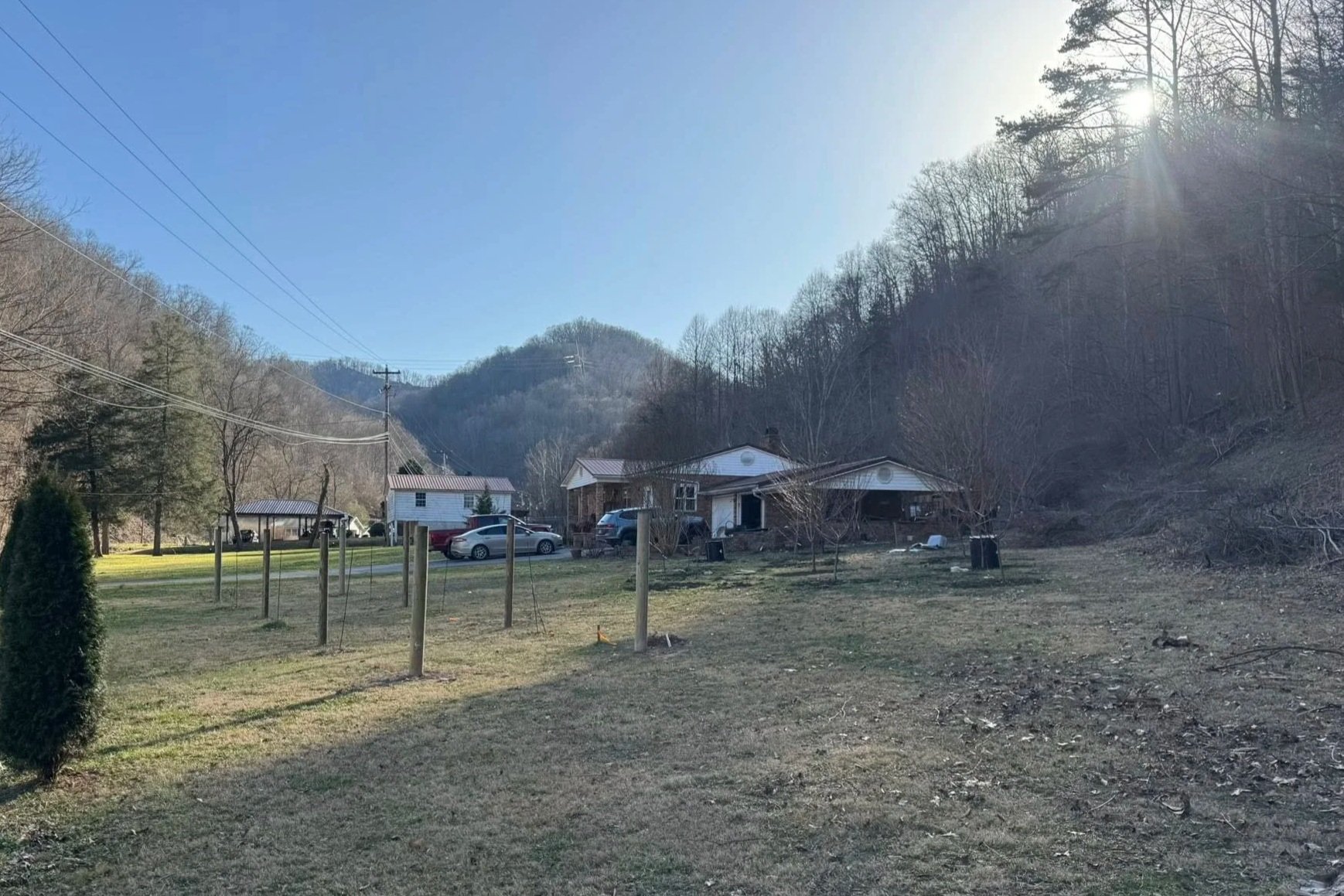 A rural scene with houses on a grassy lot, parked cars, utility poles and power lines, leafless trees, and a mountain in the background under a clear blue sky with the sun shining from the upper right corner.