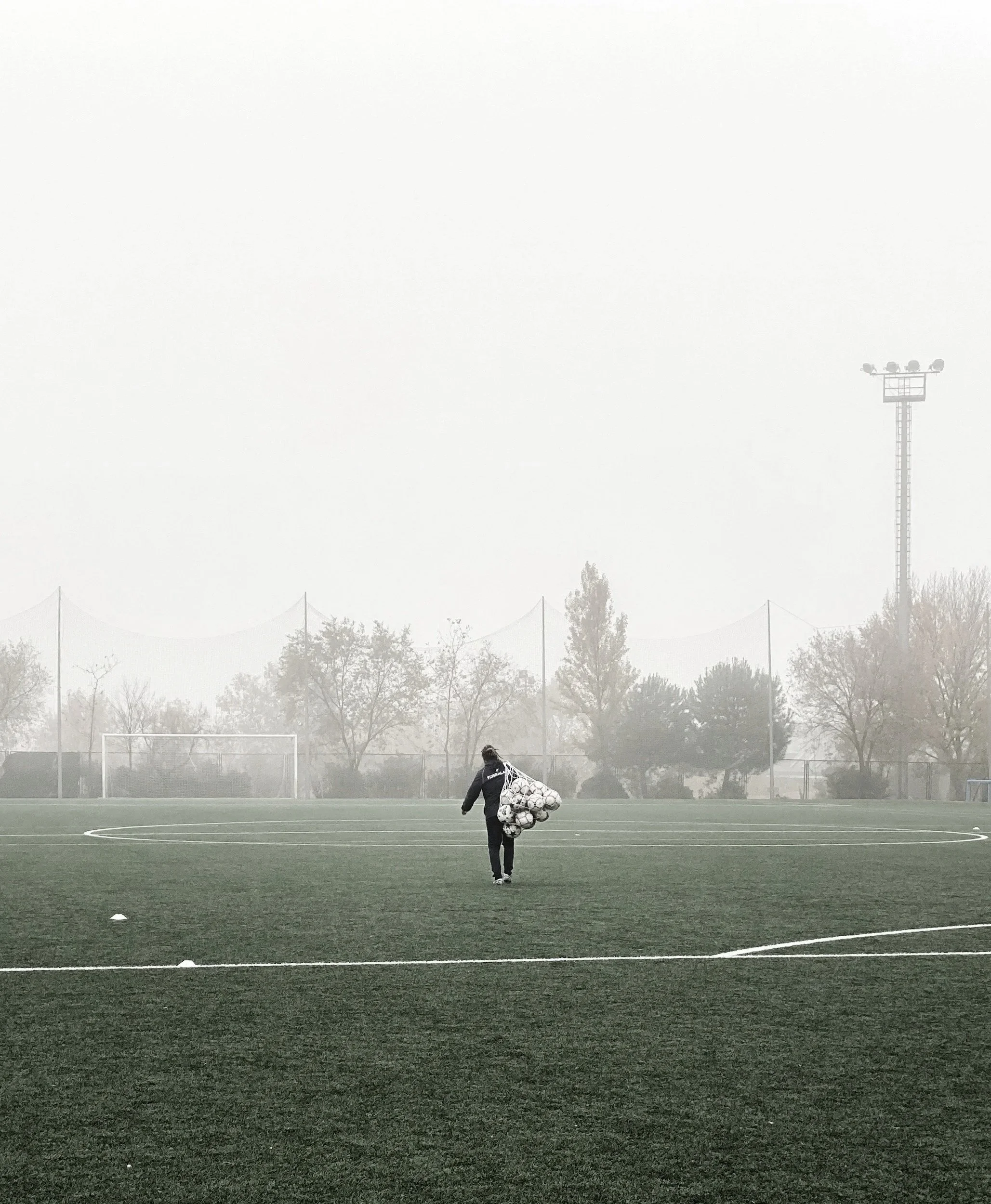 A person walking on a foggy soccer field carrying a bag filled with soccer balls.