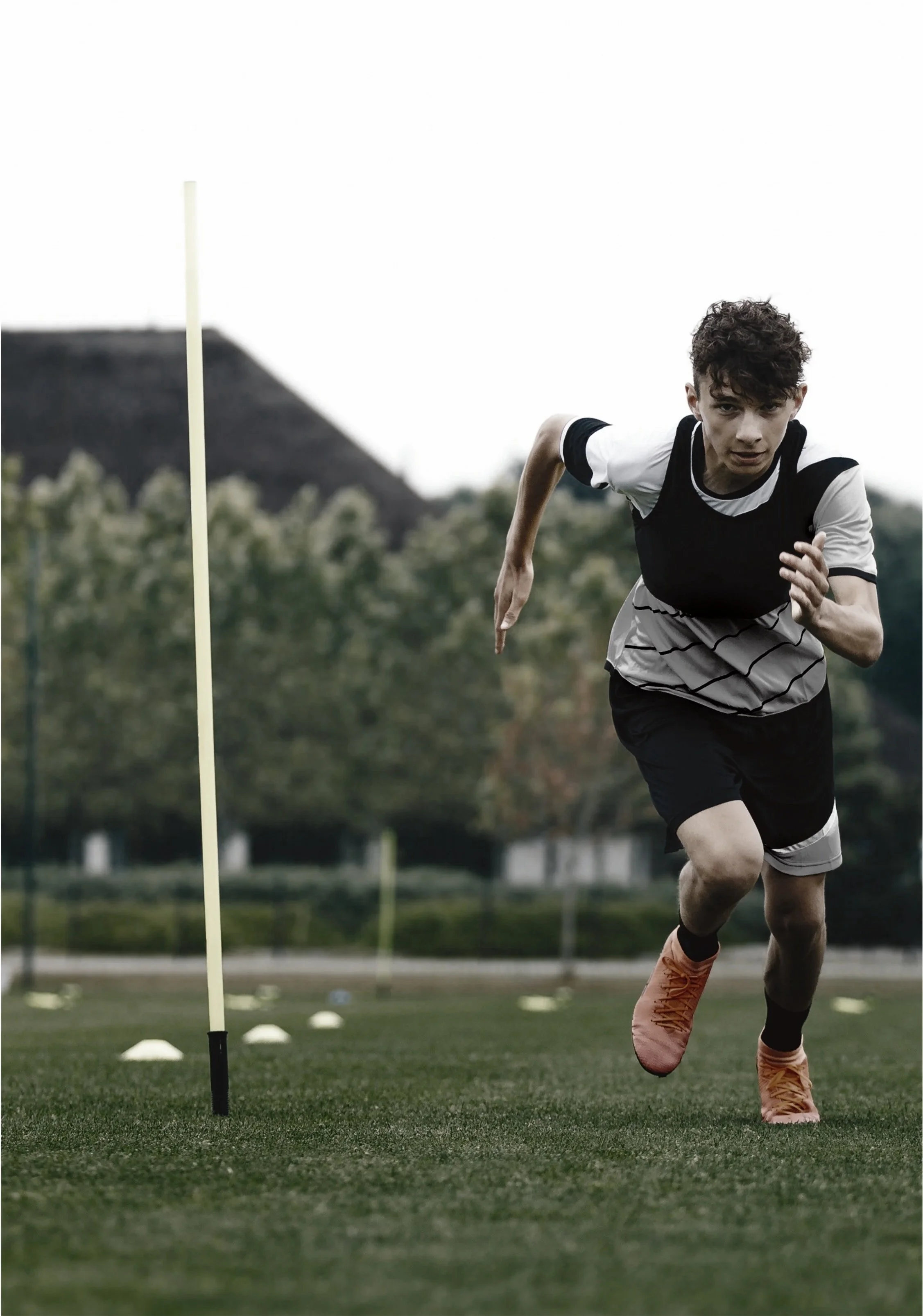 Young male athlete running on a grassy sports field during a training session with training cones and a pole in the background.