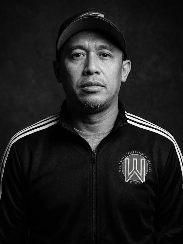 Black and white portrait of a man wearing a black cap and a black zip-up athletic jacket with white stripes and a logo on the left chest reading 'Westcalfie International Academy EST 2019'.