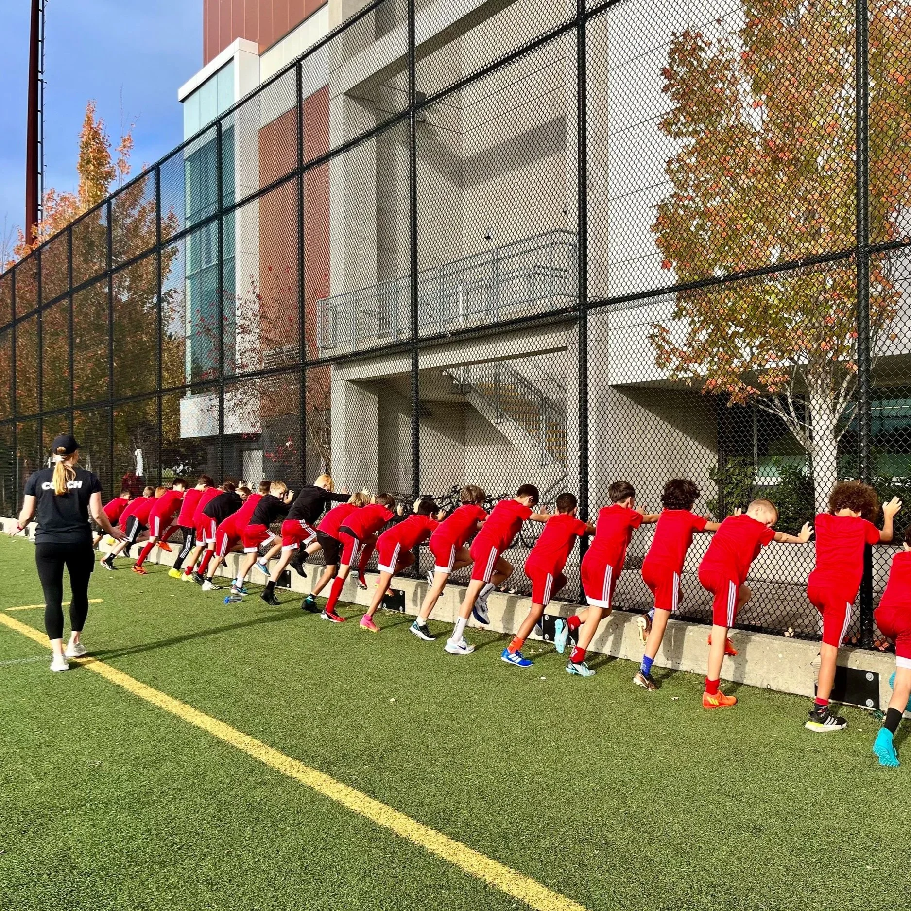 A group of young soccer players in red uniforms stretching against a black chain-link fence on a green artificial turf field, with a modern building and trees in the background. An adult woman in a black shirt with white text is overseeing them.