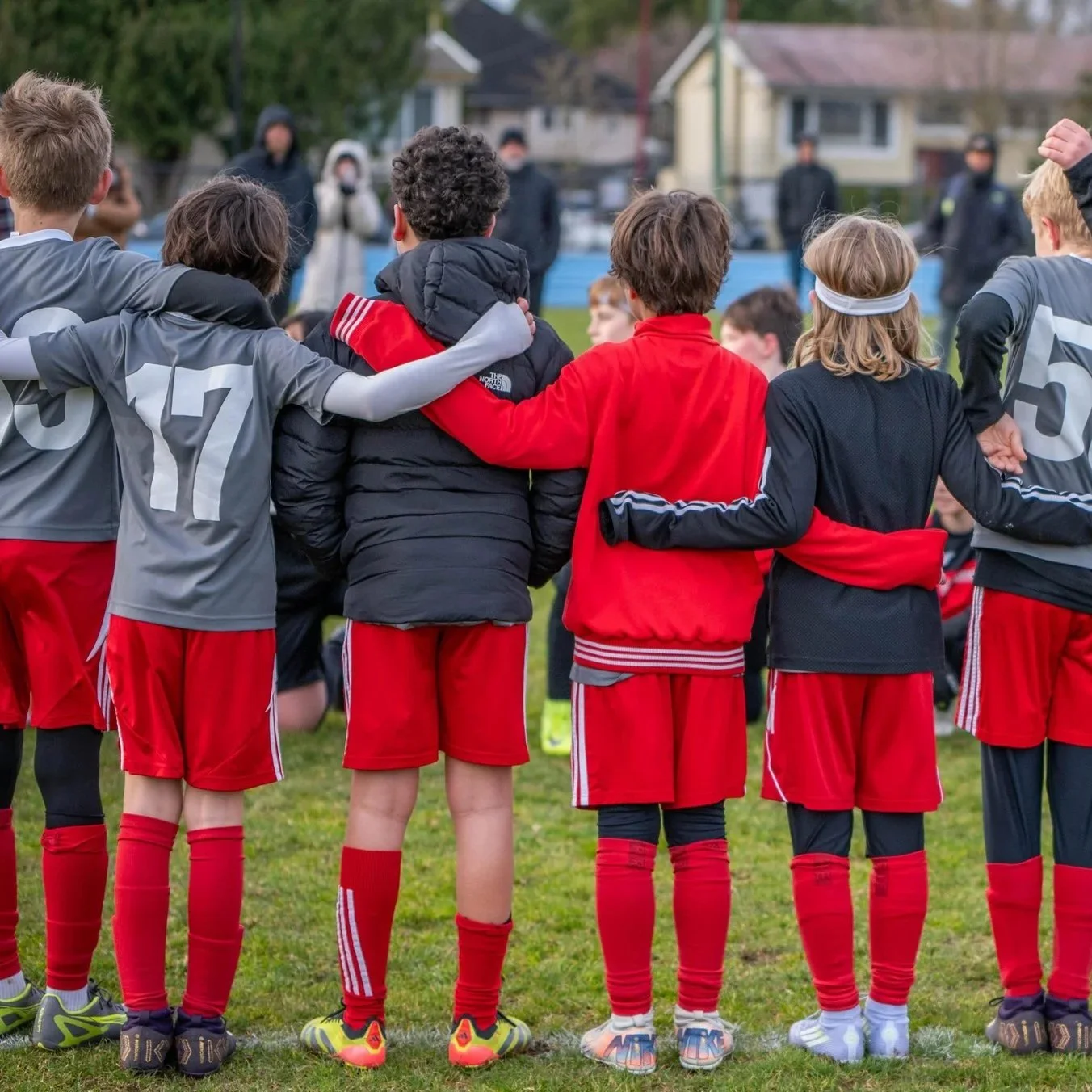 Young soccer players in red uniforms stand on the field, arms around each other, watching their opponents during a game.