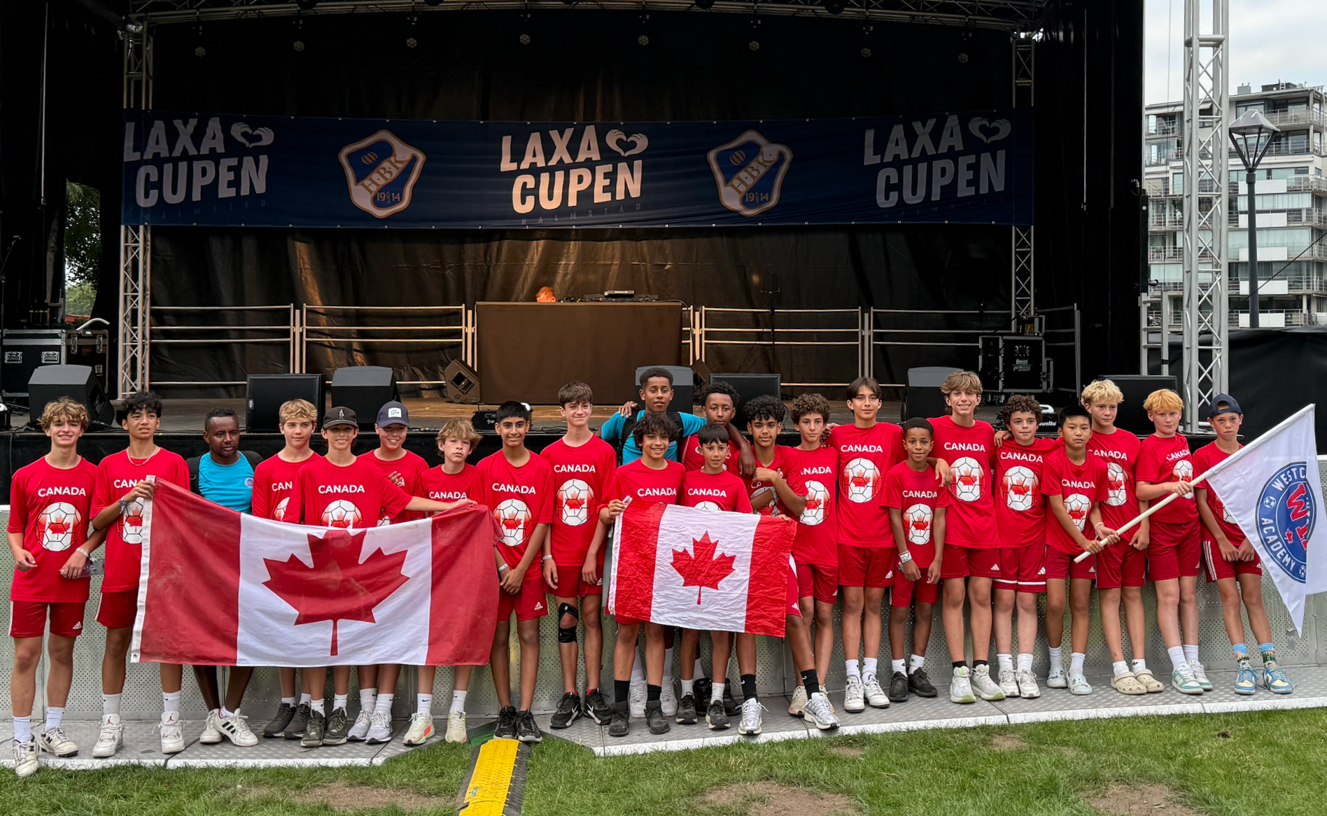 Group of young boys and girls wearing red soccer uniforms with Canadian flags and Canada printed on their shirts, standing in front of a stage with a banner that reads "LAXA CUPEN". Some are holding flags, and they are outdoors with buildings visible