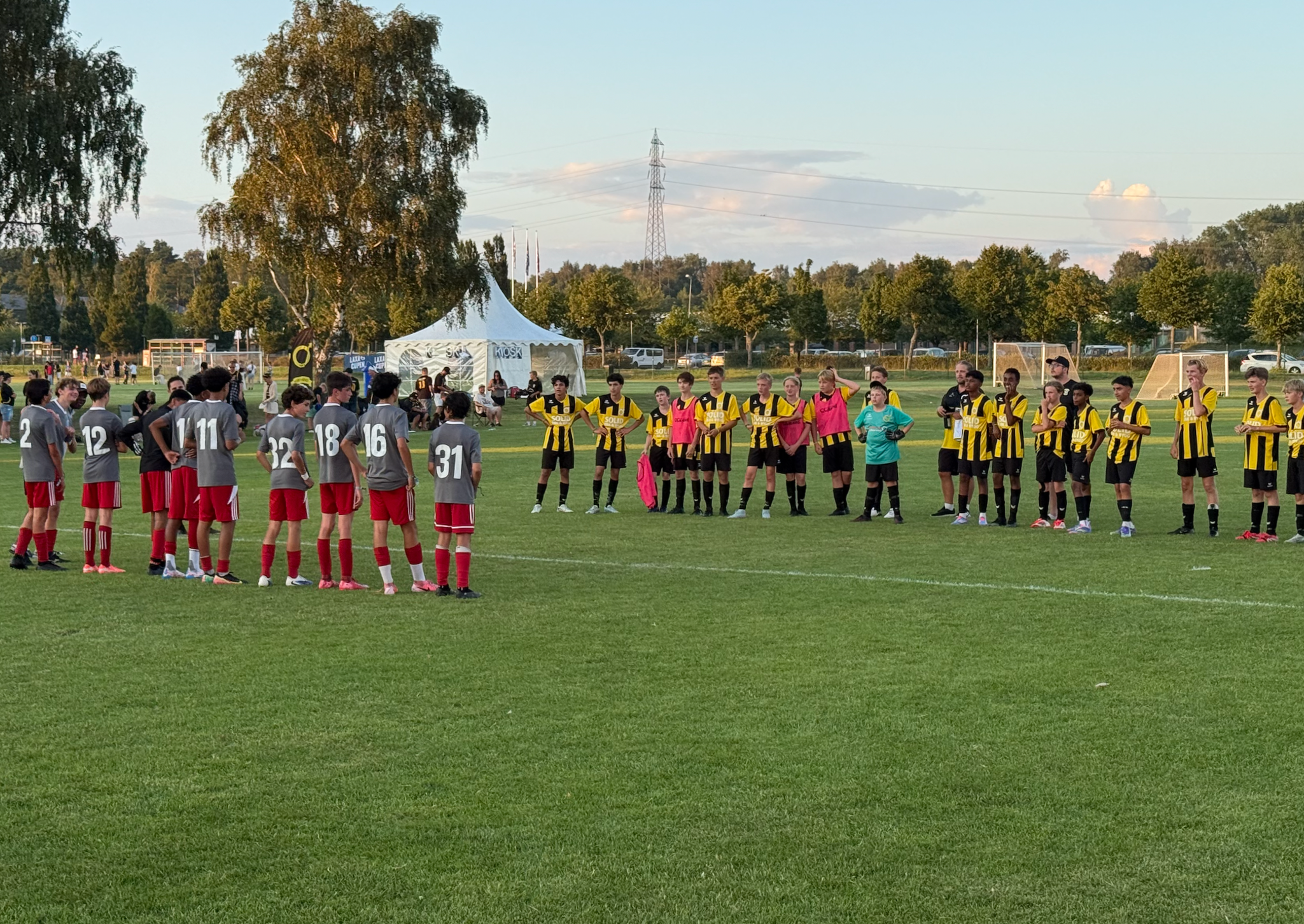 Youth soccer players standing on the field before a game, some in gray and red uniforms, others in black and yellow jerseys, with a white tent and trees in the background.
