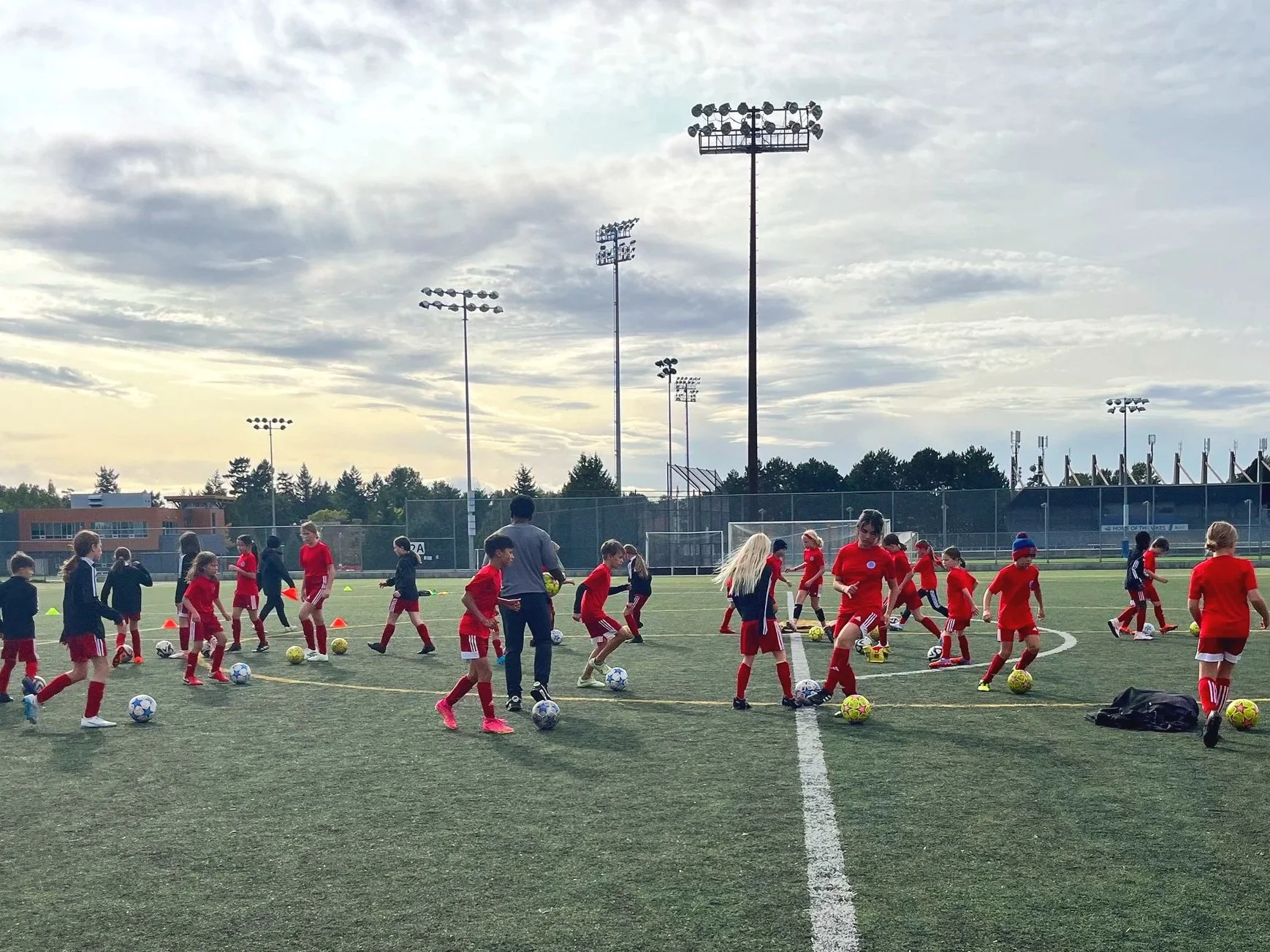 Children and coaches practicing soccer drills on a field during late afternoon. Kids are wearing red uniforms, and some are wearing athletic shoes, scattered with soccer balls and training cones. The field is outdoors with tall stadium lights and a backdrop of trees and buildings under a cloudy sky.