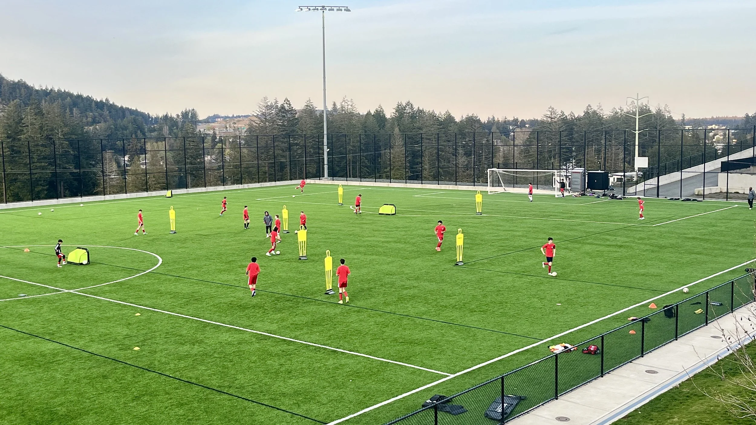 Soccer players in red jerseys practicing on a green outdoor field with training mannequins and cones, surrounded by a black fence, with a backdrop of trees and hills.