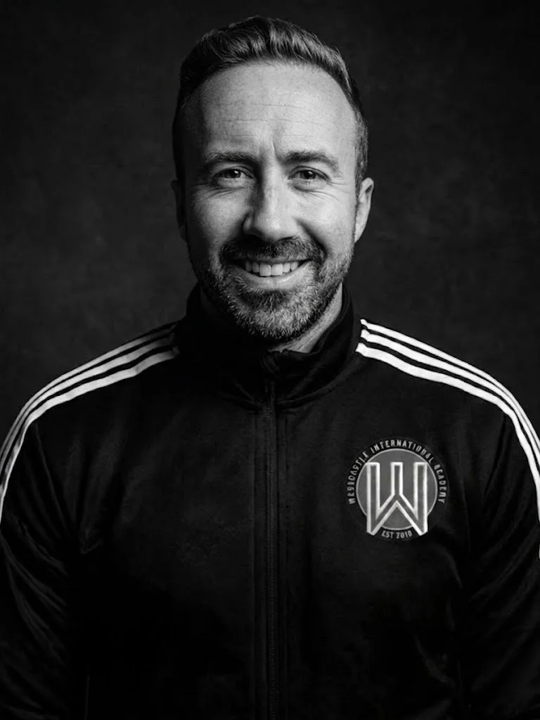Black and white portrait of a smiling man with short hair and a beard, wearing a sports jacket with stripes on the shoulders and a logo on the chest that says "MissionFit International Club".