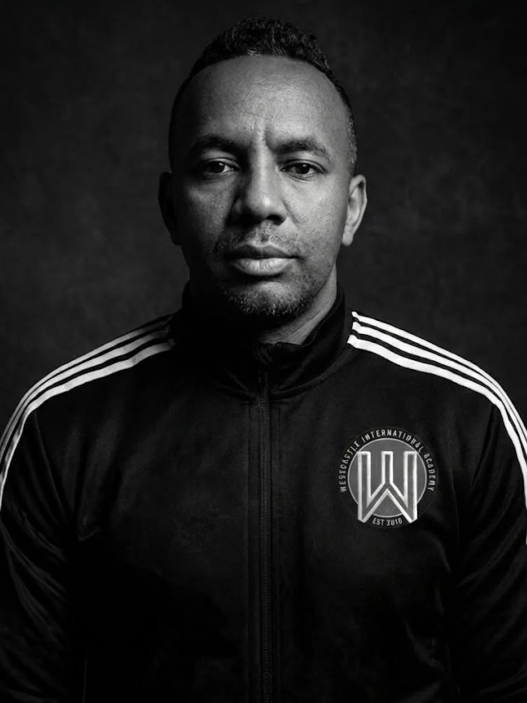 Black and white portrait of a man wearing a sports jacket with the logo and text 'Wesleyan International Academy' on the chest, and white stripes on the shoulders.