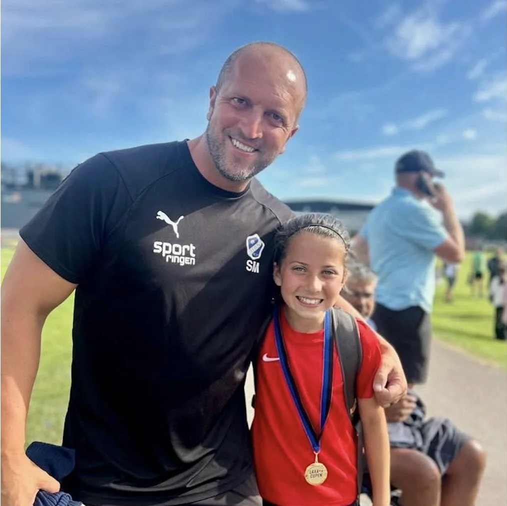 A man and girl smiling outdoors after a sports event. The man is wearing a black sports shirt with Puma logo and is muscular with a beard. The girl is wearing a red sports shirt with a medal around her neck and has a backpack. The background shows ot