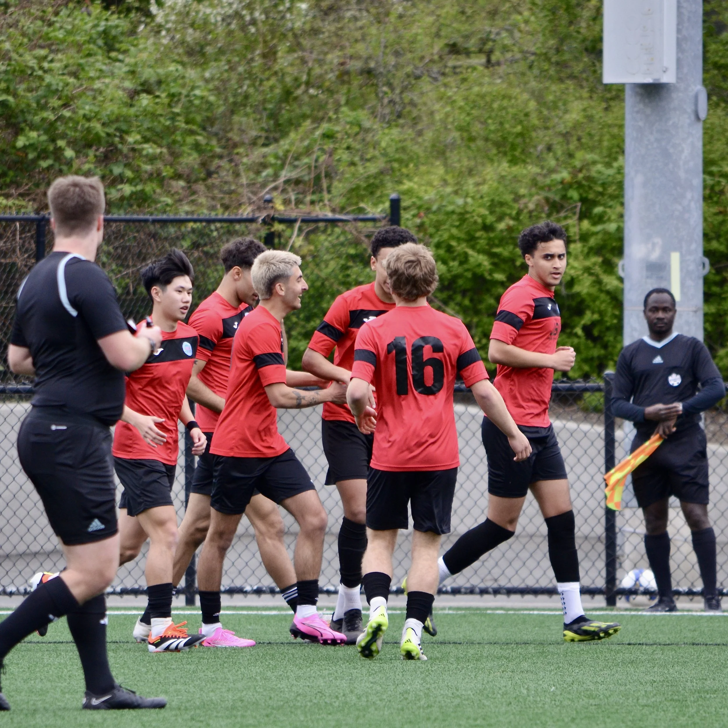 A group of soccer players wearing red jerseys and black shorts on the field during a game. Some players have blonde and dark hair. One player has a visible number 10 on his jersey. The background shows a chain-link fence and green trees.