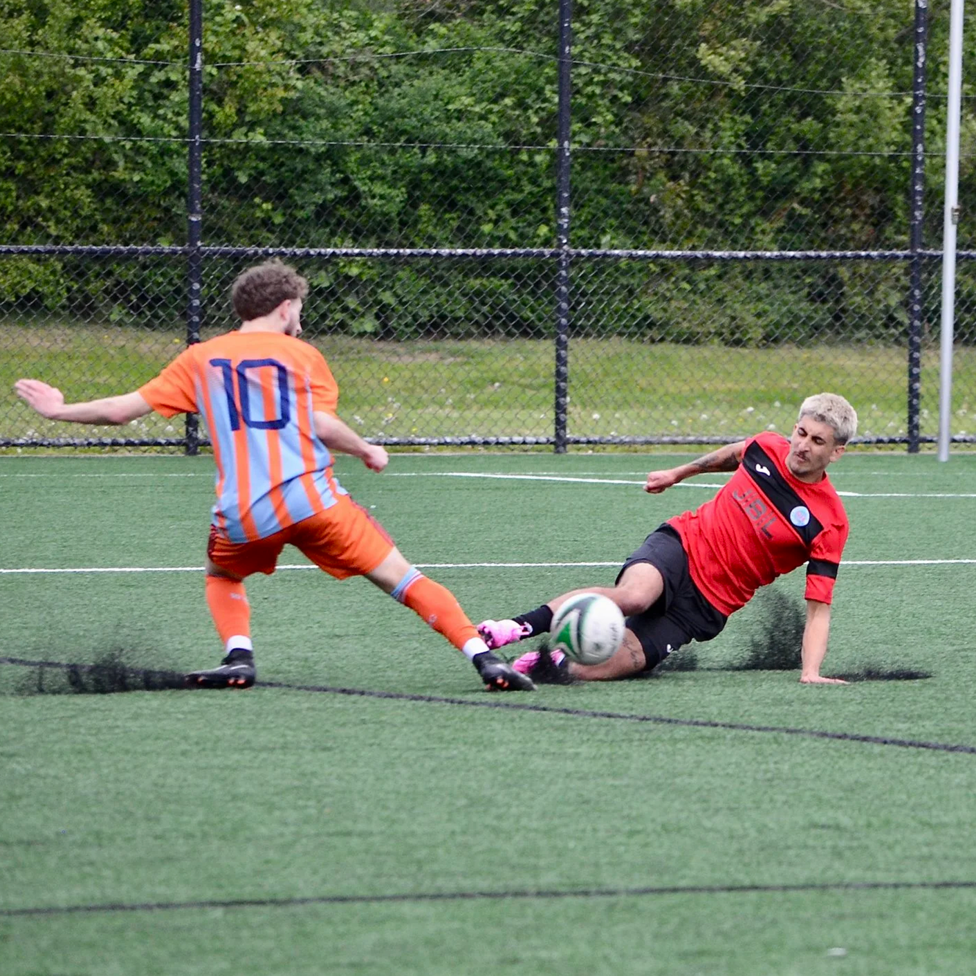 A soccer player in an orange and blue striped jersey tries to kick the ball while a player in a red jersey slides on the ground to block the shot during a soccer match on a green field with a black fence and trees in the background.