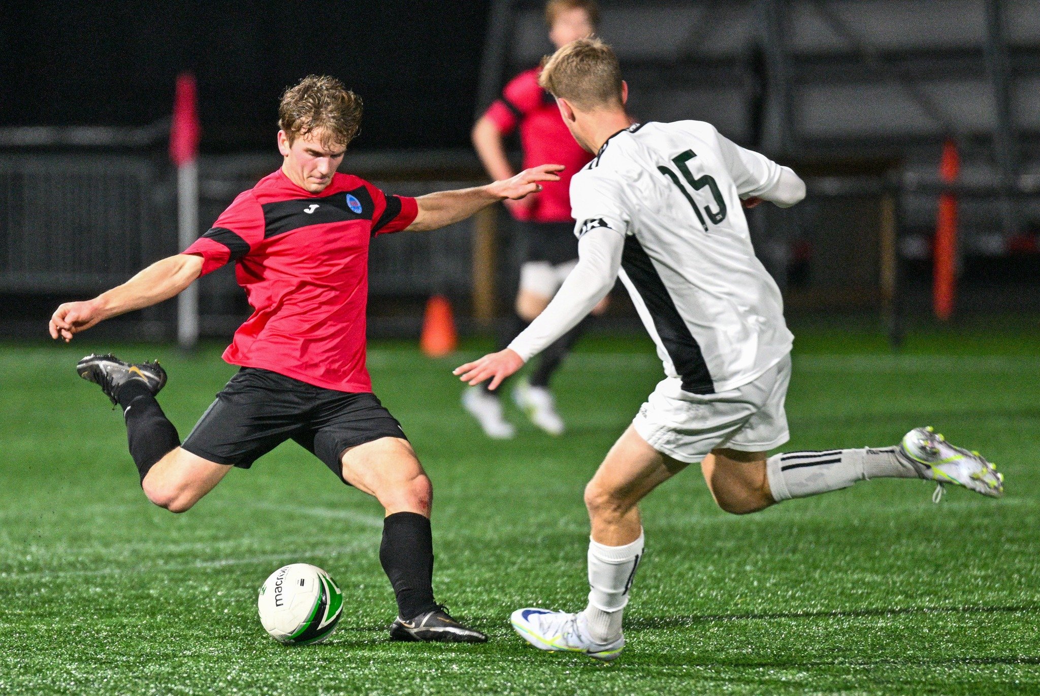 Two male soccer players competing for the ball on an outdoor field at night. One is wearing a red and black jersey, and the other is wearing a white jersey with the number 15. The player in red is kicking the ball while the player in white attempts to block.