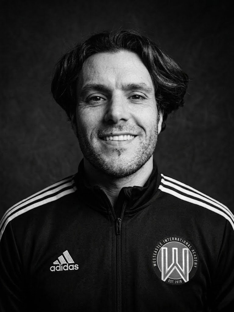Black and white photo of a smiling man wearing a sports jacket with Adidas logo and Westside International Academy badge.