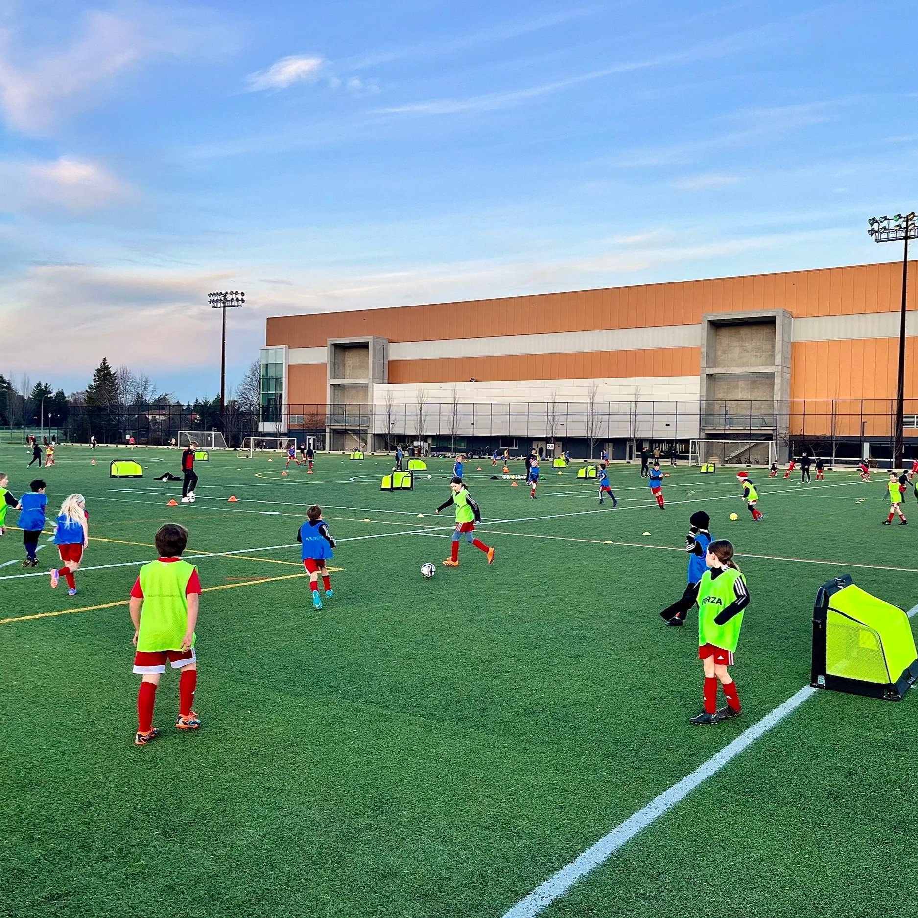 Children playing soccer on a green field during daytime, some wearing yellow bibs, with a large orange and gray building, tall floodlights, and trees in the background, under a mostly clear sky with scattered clouds.