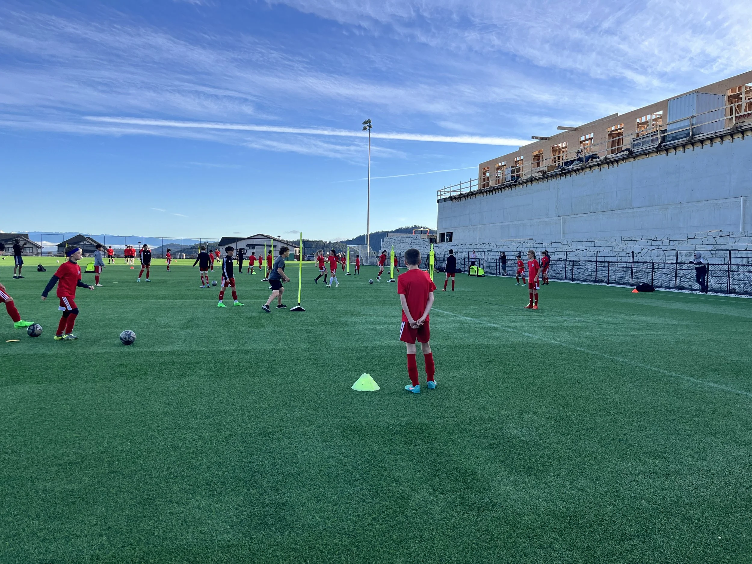 Soccer players practicing drills on a green field, with a construction site and blue sky in the background.