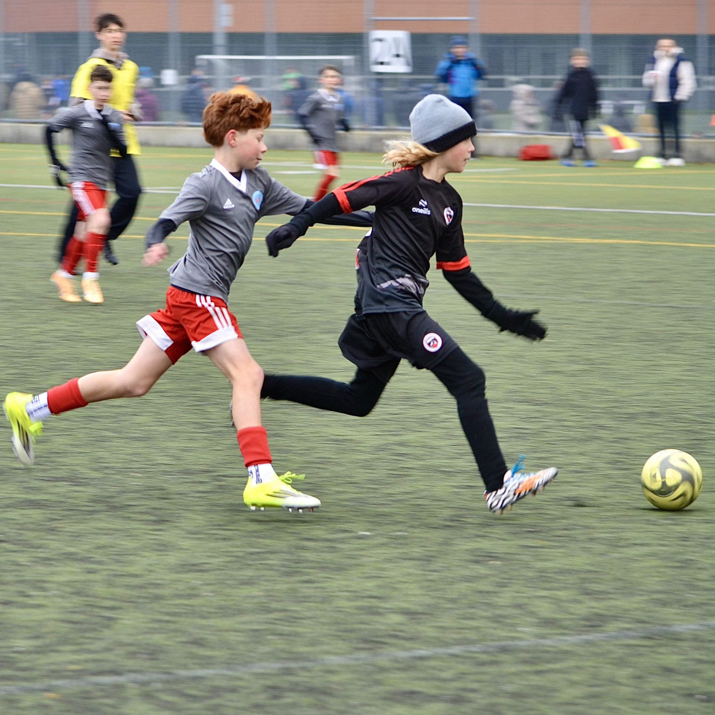 Children playing soccer on a field, with others watching in the background.