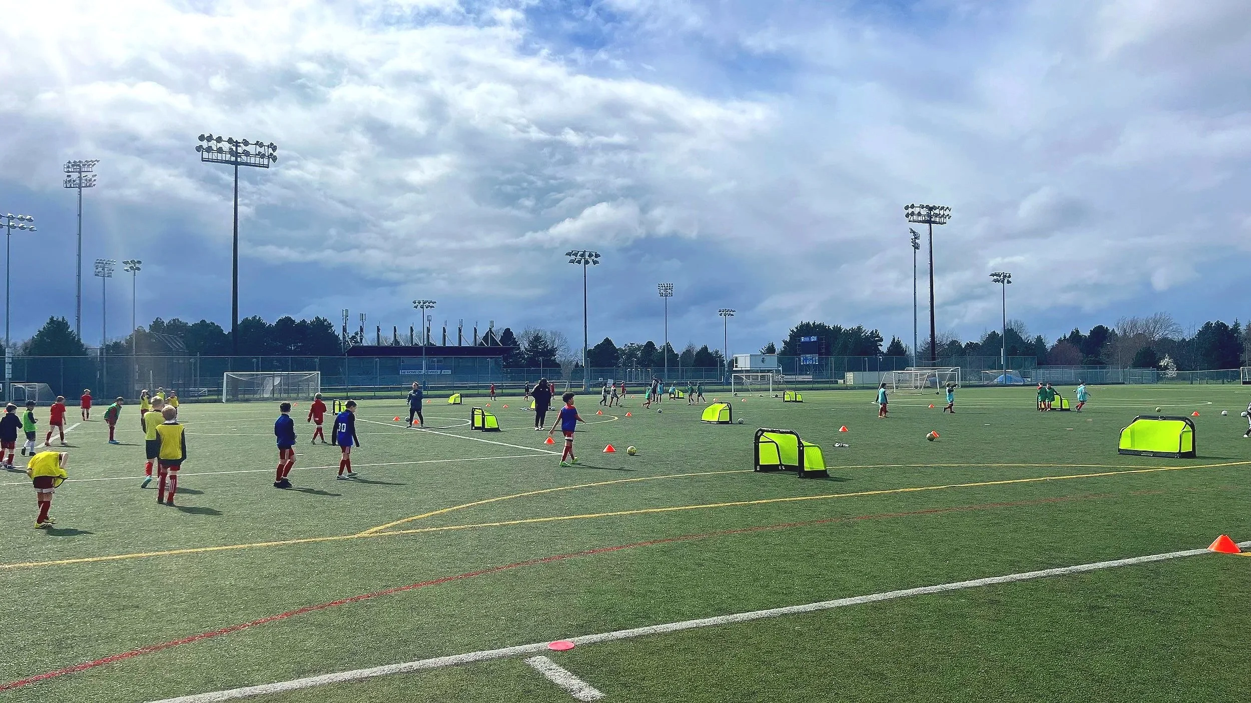 Young children participating in a soccer practice on a large outdoor field with multiple goals and training equipment. Overcast sky with some blue patches and tall stadium lights.
