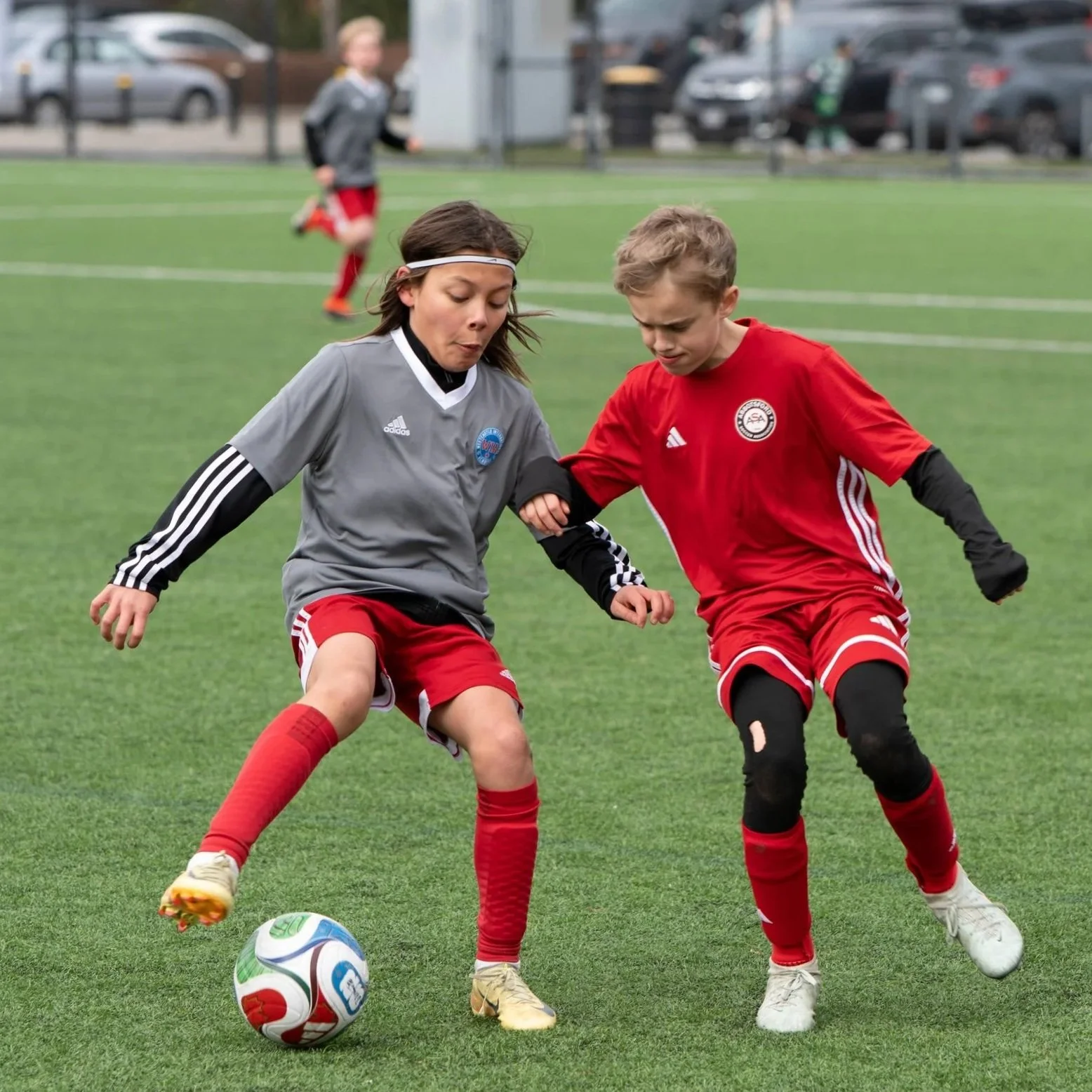 Two young children playing soccer on a field, both wearing red shorts and soccer jerseys, challenging for the ball, with a pubic soccer ball in front of them.