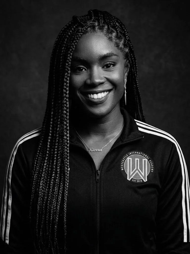 Black and white portrait of a smiling young woman with long braided hair, wearing a sports jacket with a badge from Westside International Academy and a necklace with the name 'Shenqa'.