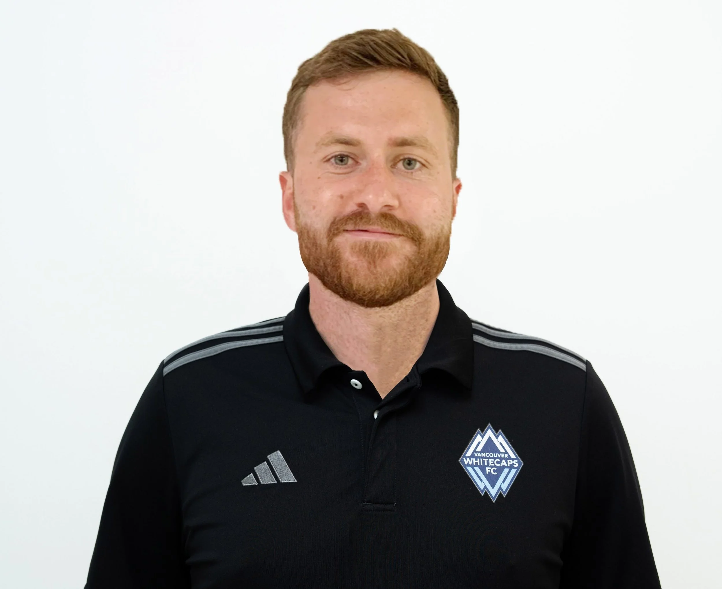 A man with light skin, short red hair, and a beard wearing a Vancouver Whitecaps FC jacket, standing in front of a plain white background.
