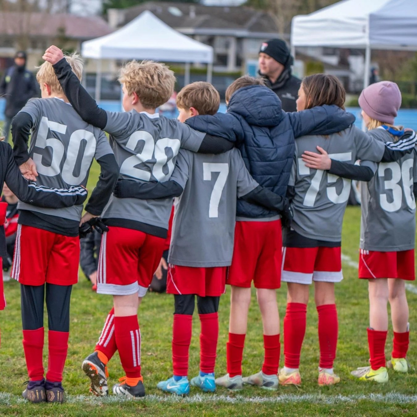 Young soccer players in gray jerseys with red shorts, standing in a line with arms around each other's shoulders, on a grassy field during daytime.