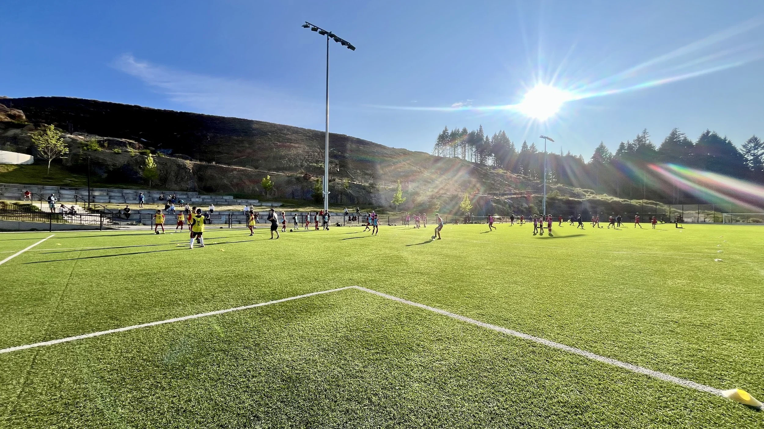 Soccer game being played on a sunny day on a green field with players, some in yellow bibs, and spectators on bleachers, surrounded by hills and trees.