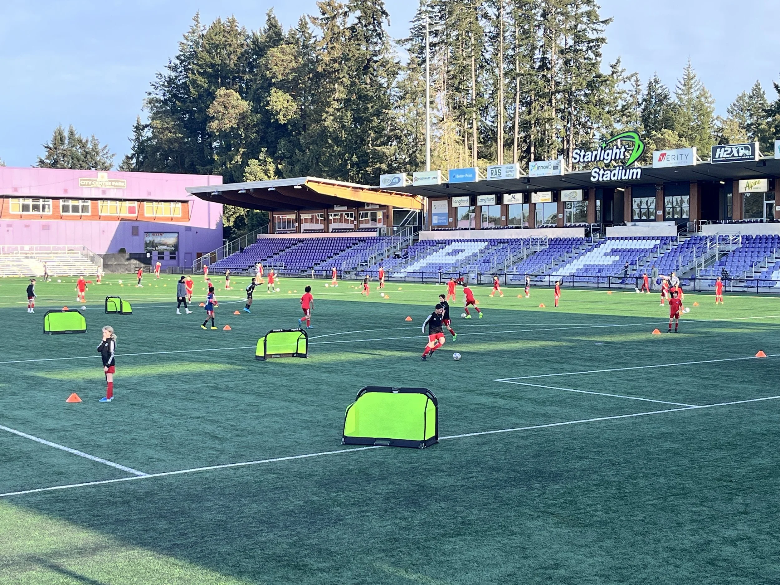 A soccer training session taking place on a large outdoor field, with children practicing drills and drills cones, in front of a stadium with purple seats and a row of trees in the background.
