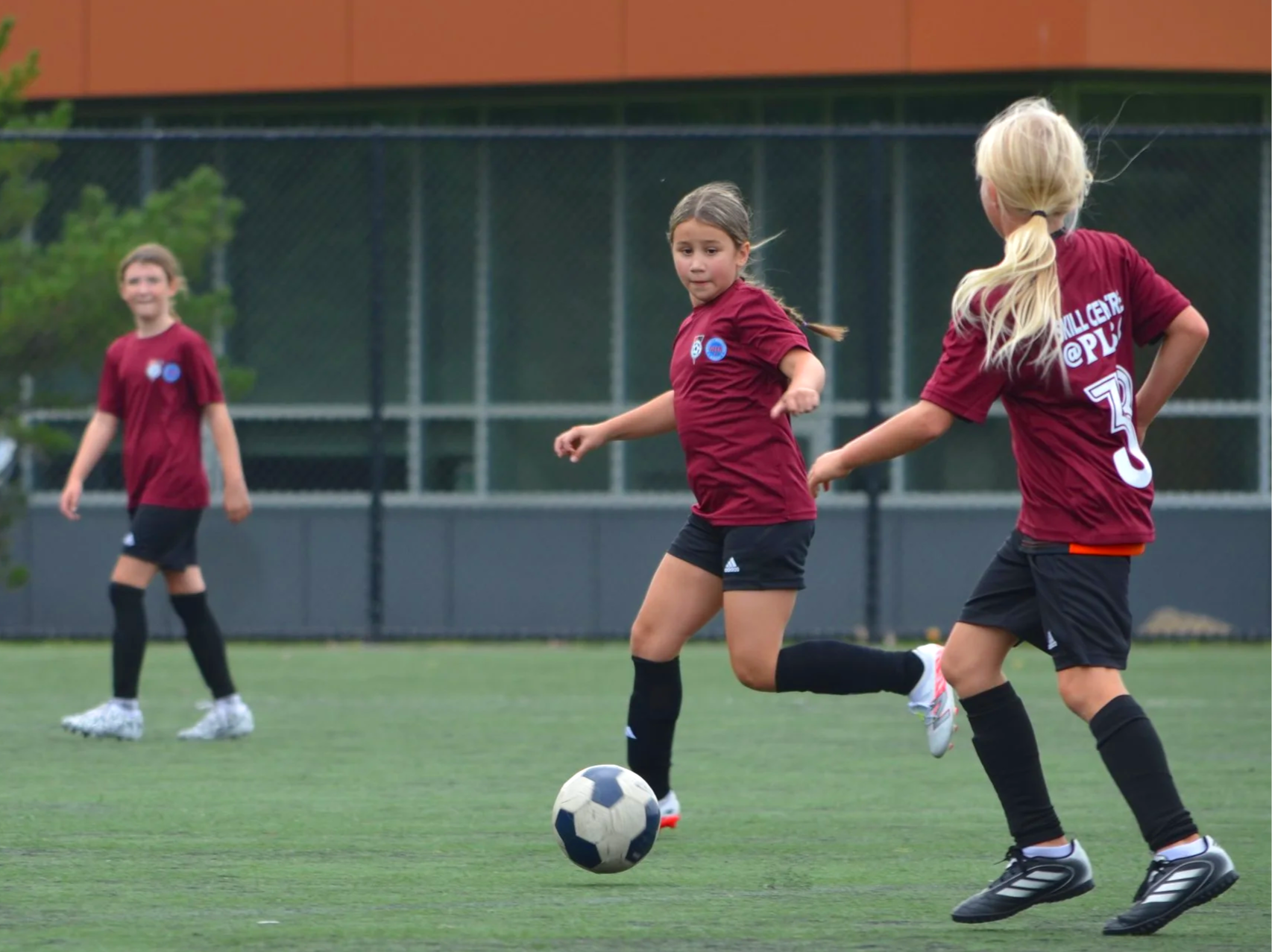 Young girls playing soccer on a field, one kicking a ball while another watches.
