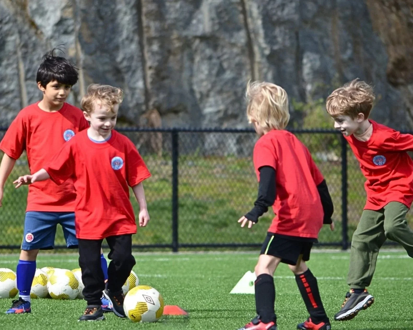 Four young boys wearing red soccer shirts practicing on a field with soccer balls and cones, with a rocky background and a chain-link fence.