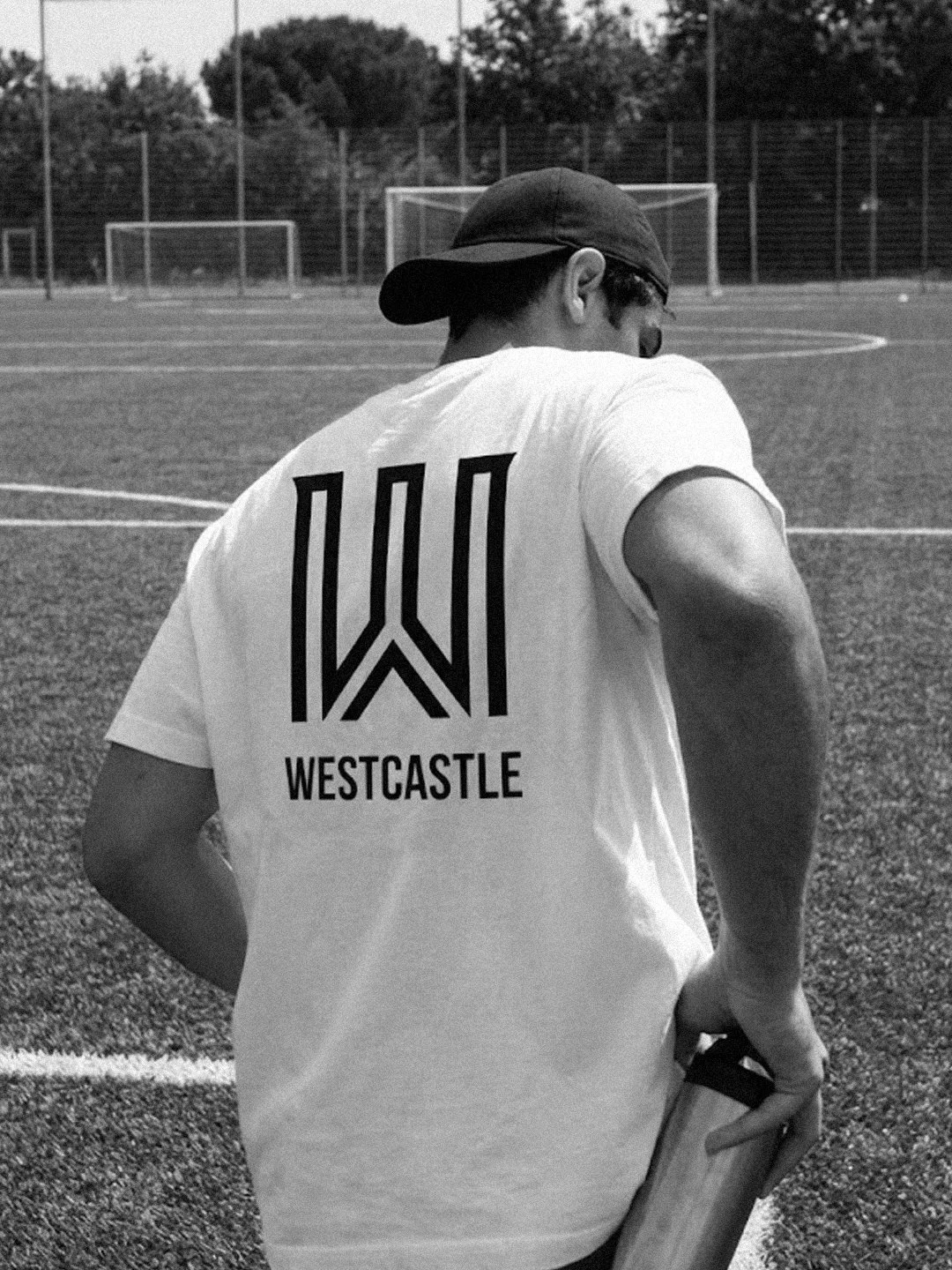 A person wearing a dark baseball cap and sunglasses, standing on a sports field, holding a water bottle, with their back to the camera. They are wearing a t-shirt with the word 'Westcastle' and a geometric design on the back.