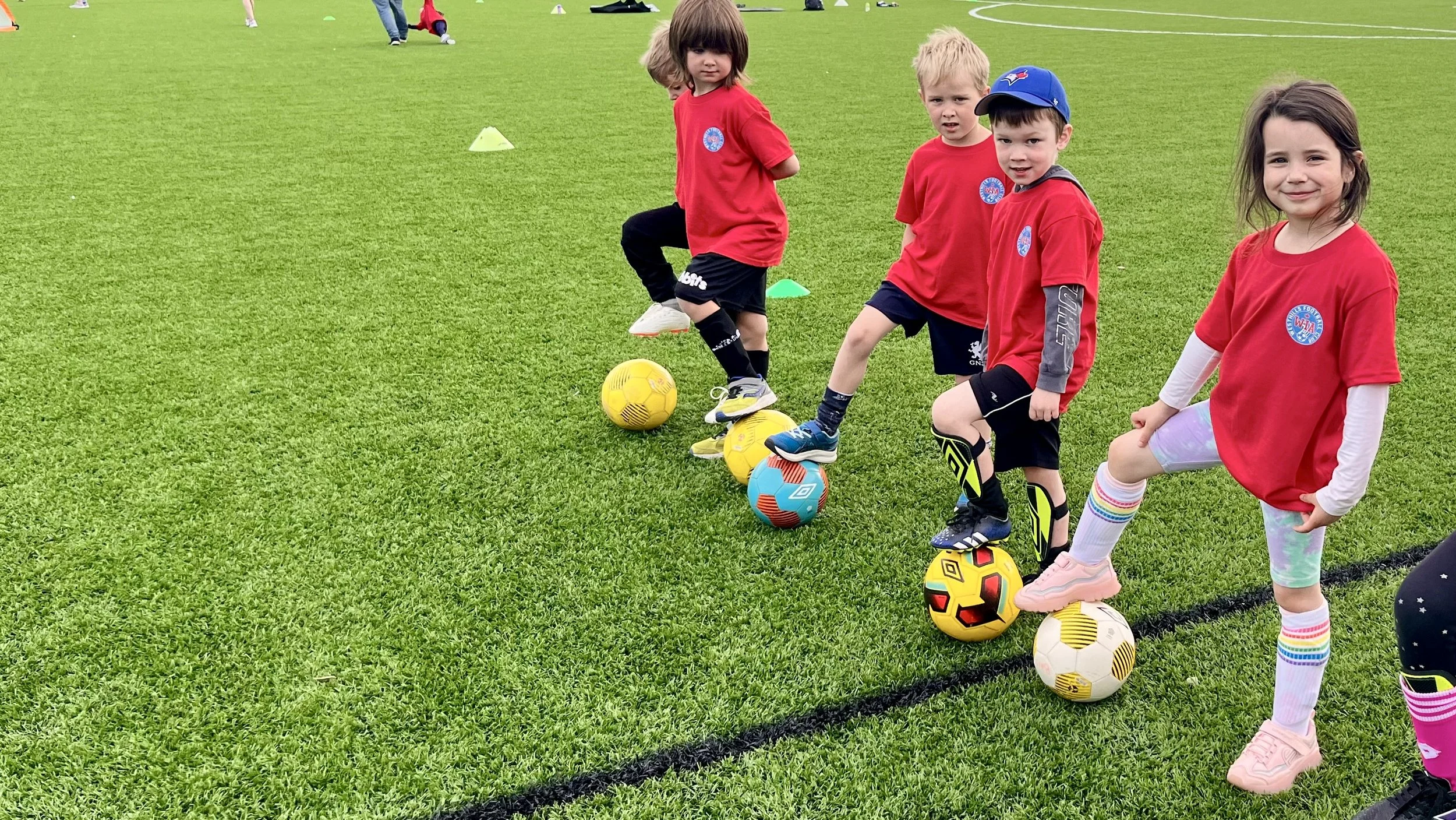A group of young children in red shirts participating in a soccer practice on a grassy field, with soccer balls lined up in front of them.