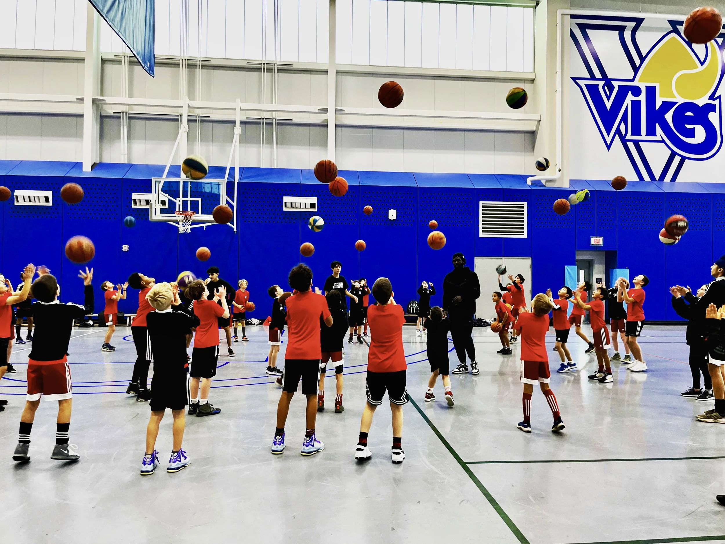 Children playing basketball indoors, some trying to catch basketballs falling from above, with a basketball hoop and logo on the wall in the background.
