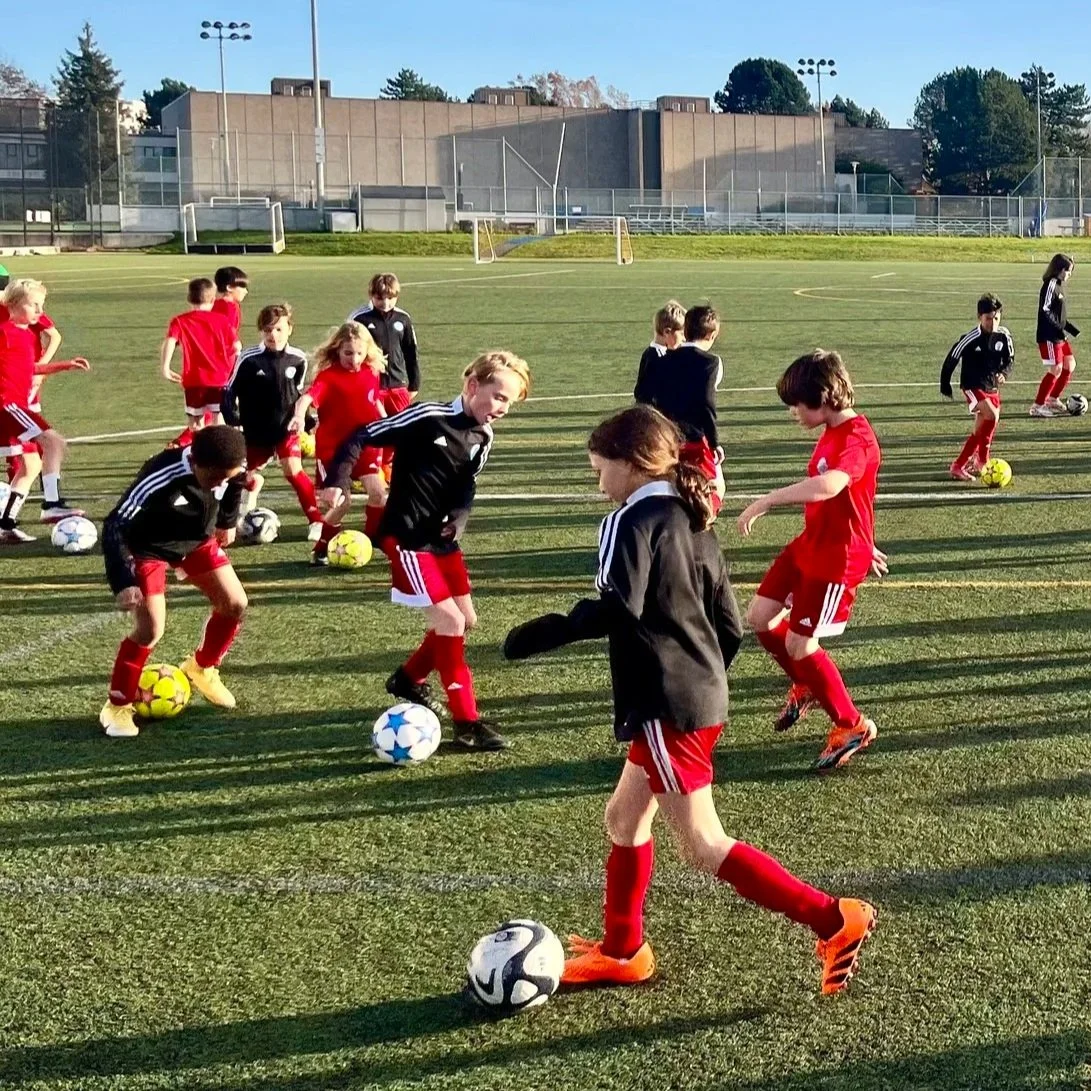 Children playing soccer on a grass field, wearing sports uniforms with cleats, with one child in red shorts and a yellow jersey kicking a soccer ball.