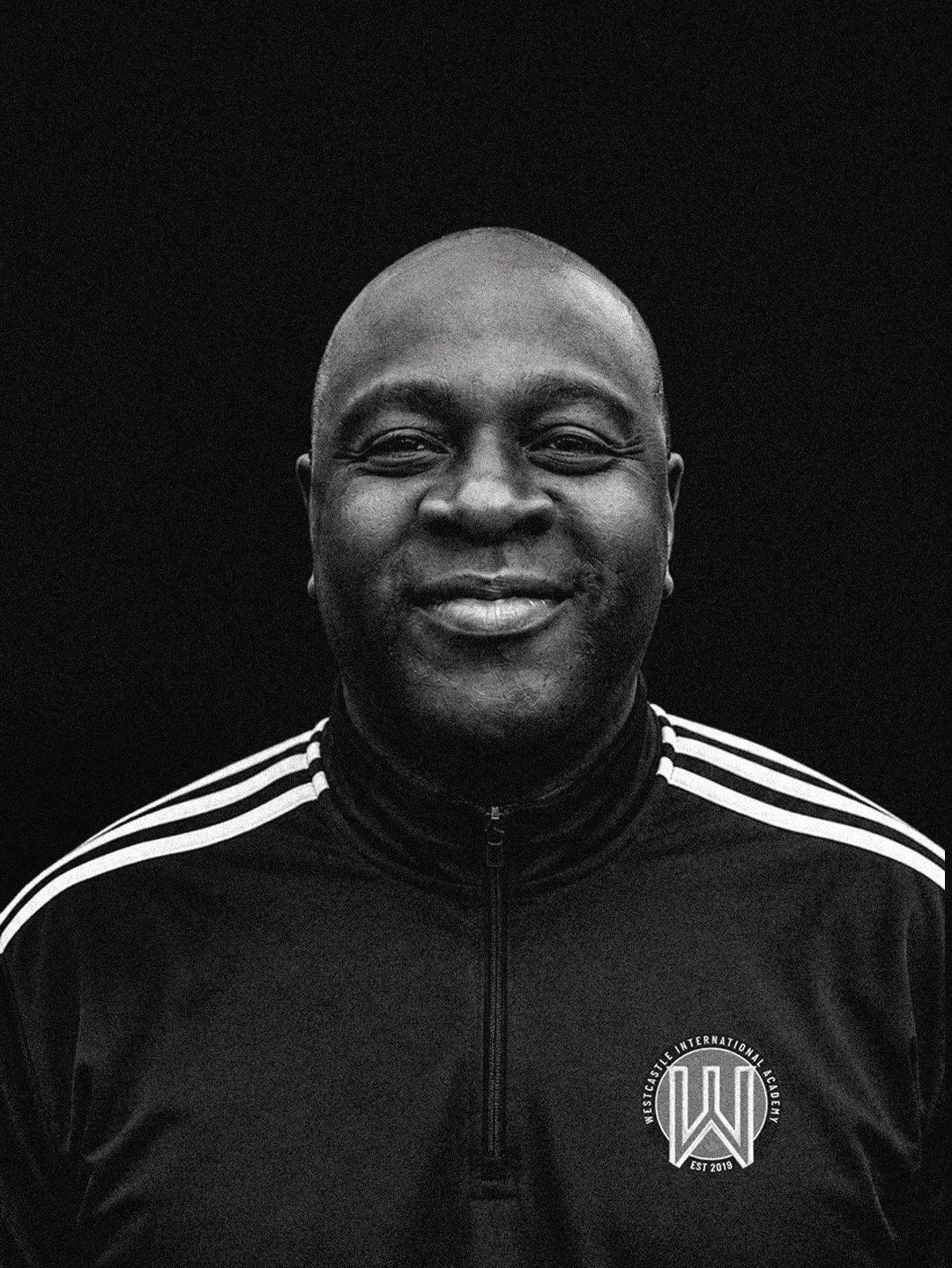 Black and white portrait of a smiling man wearing a Westcastle International Academy sports jacket with striped sleeves and a logo on the chest.