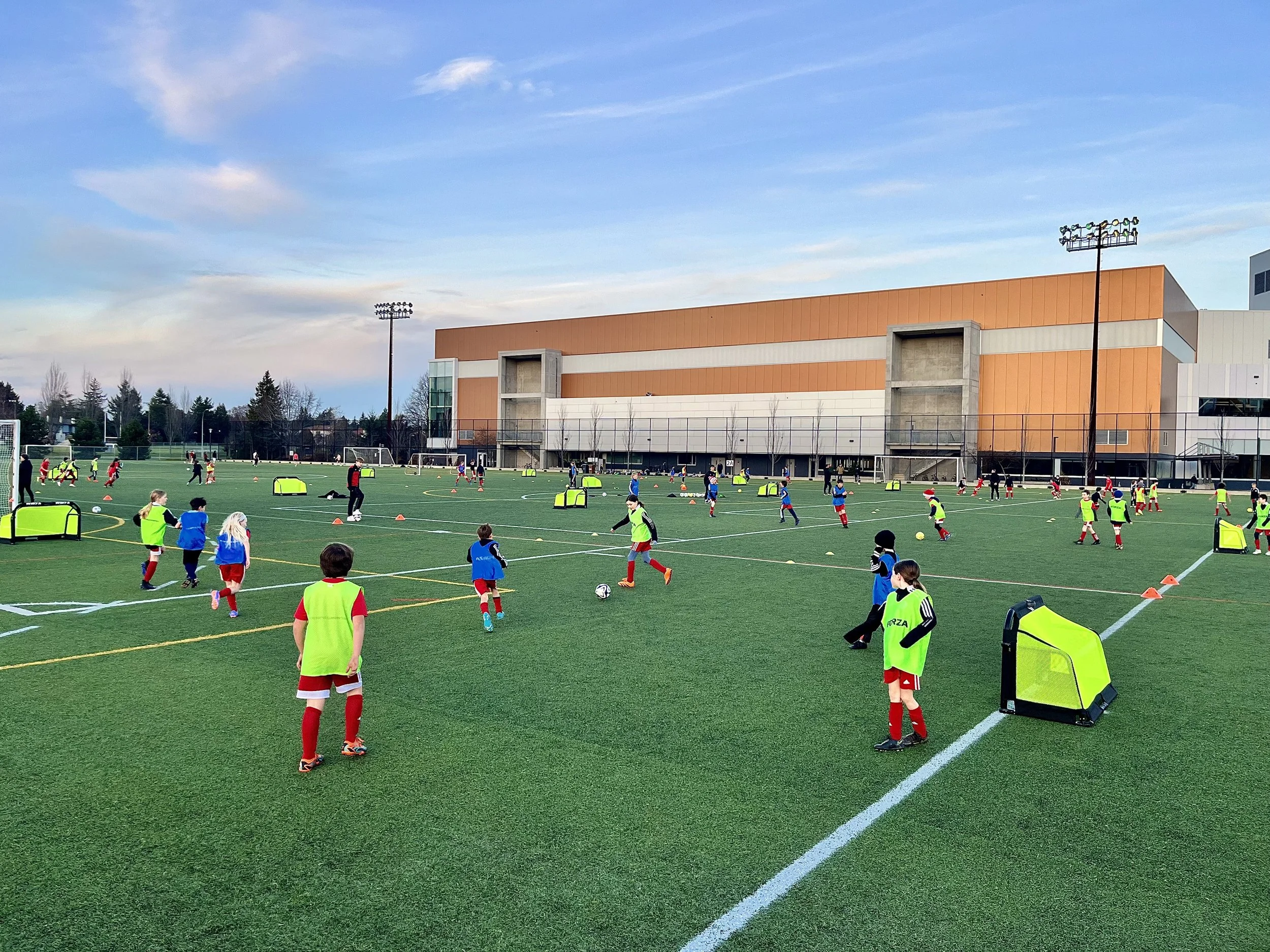 Children playing soccer on a field during practice or game, with a large brown building and trees in the background under a blue sky.