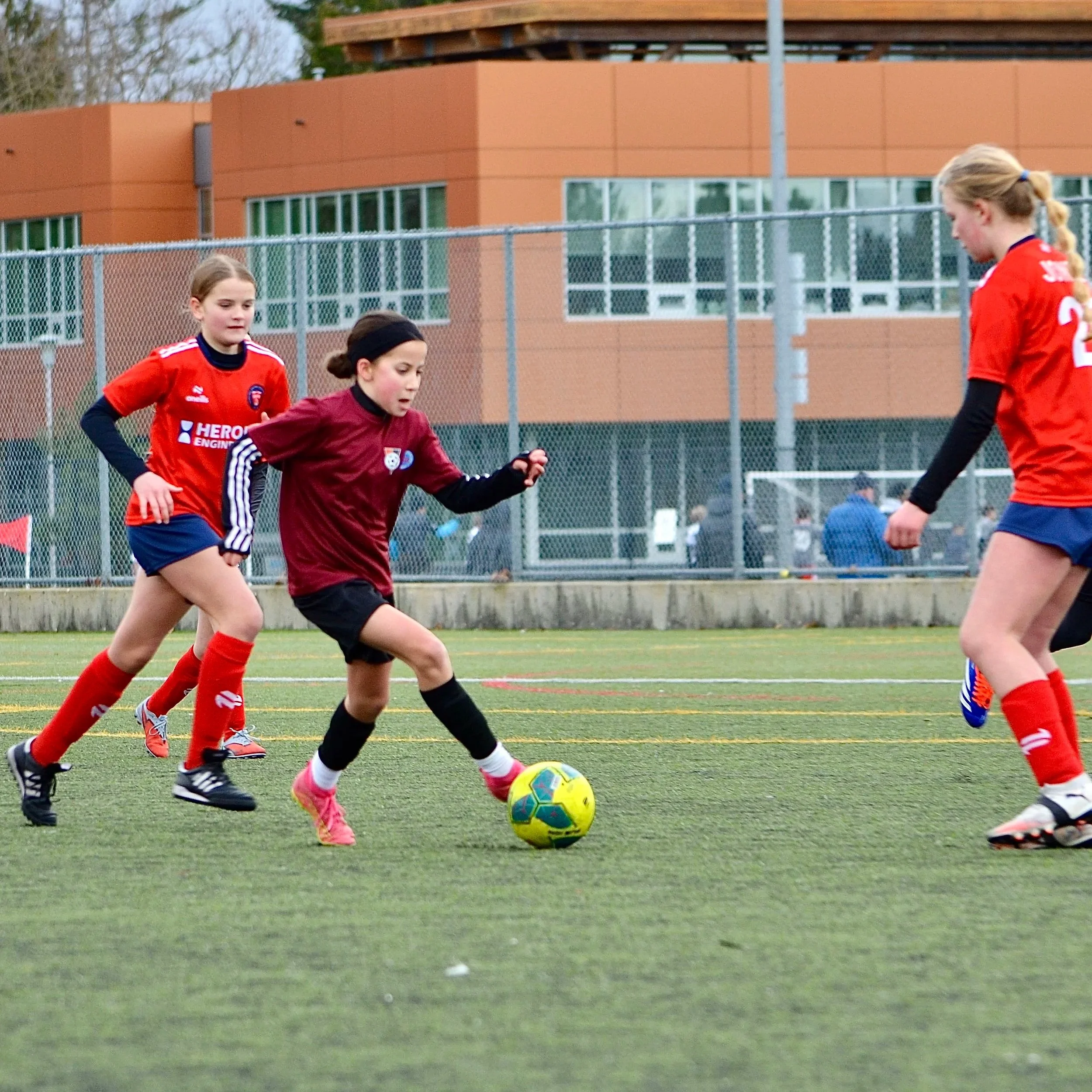 Three young girls playing soccer on an outdoor field, two girls in red jerseys and one girl in a maroon jersey, all focused on the ball.
