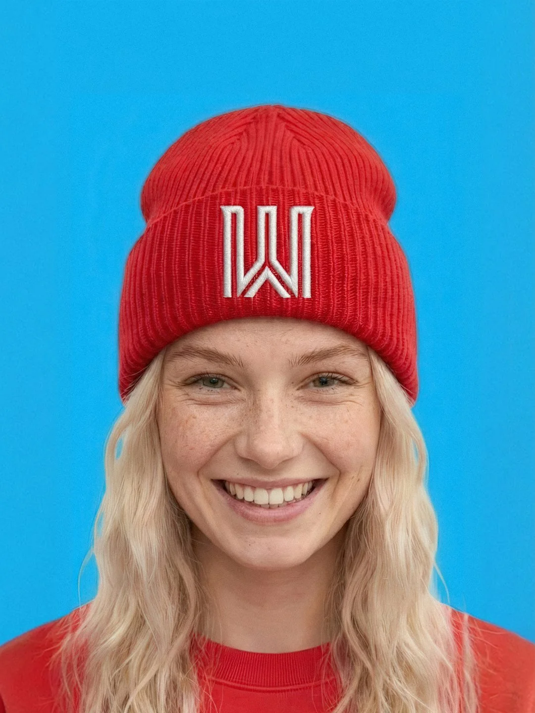 A young woman with long blonde hair wearing a red beanie with a white logo and a red shirt, smiling in front of a blue background.