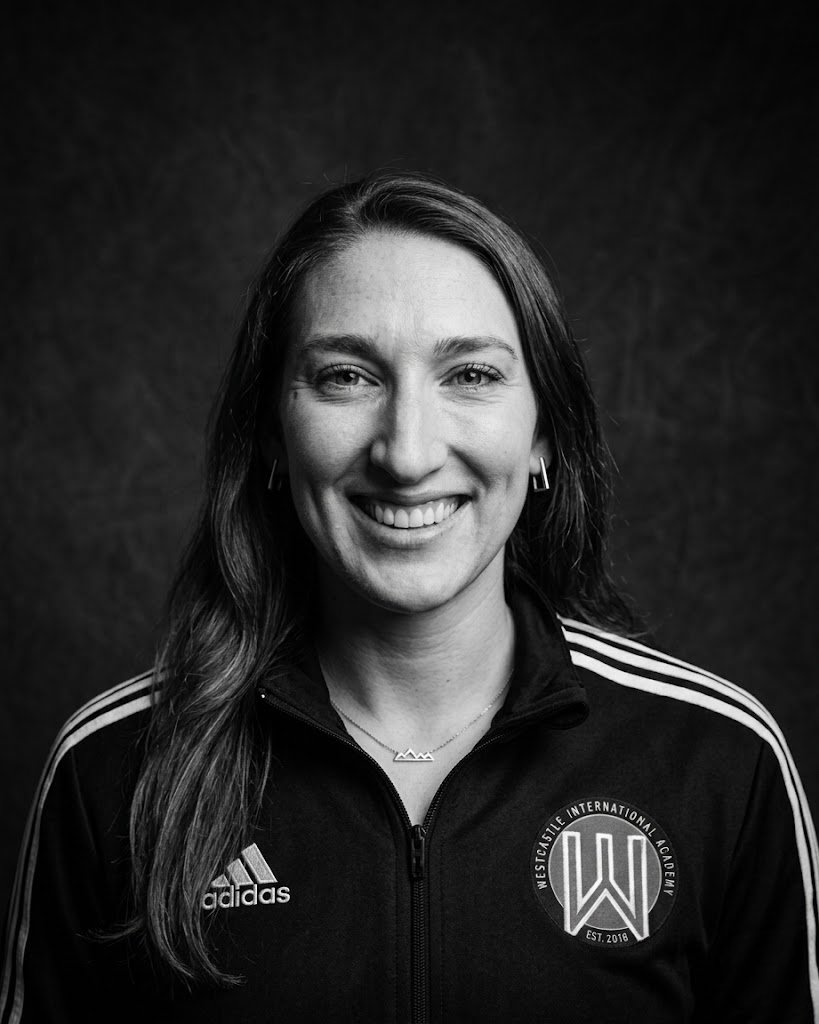 Black and white portrait photo of a smiling woman with long hair, wearing a Westcastle International Academy sports jacket with Adidas logo, and jewelry including earrings and a necklace.