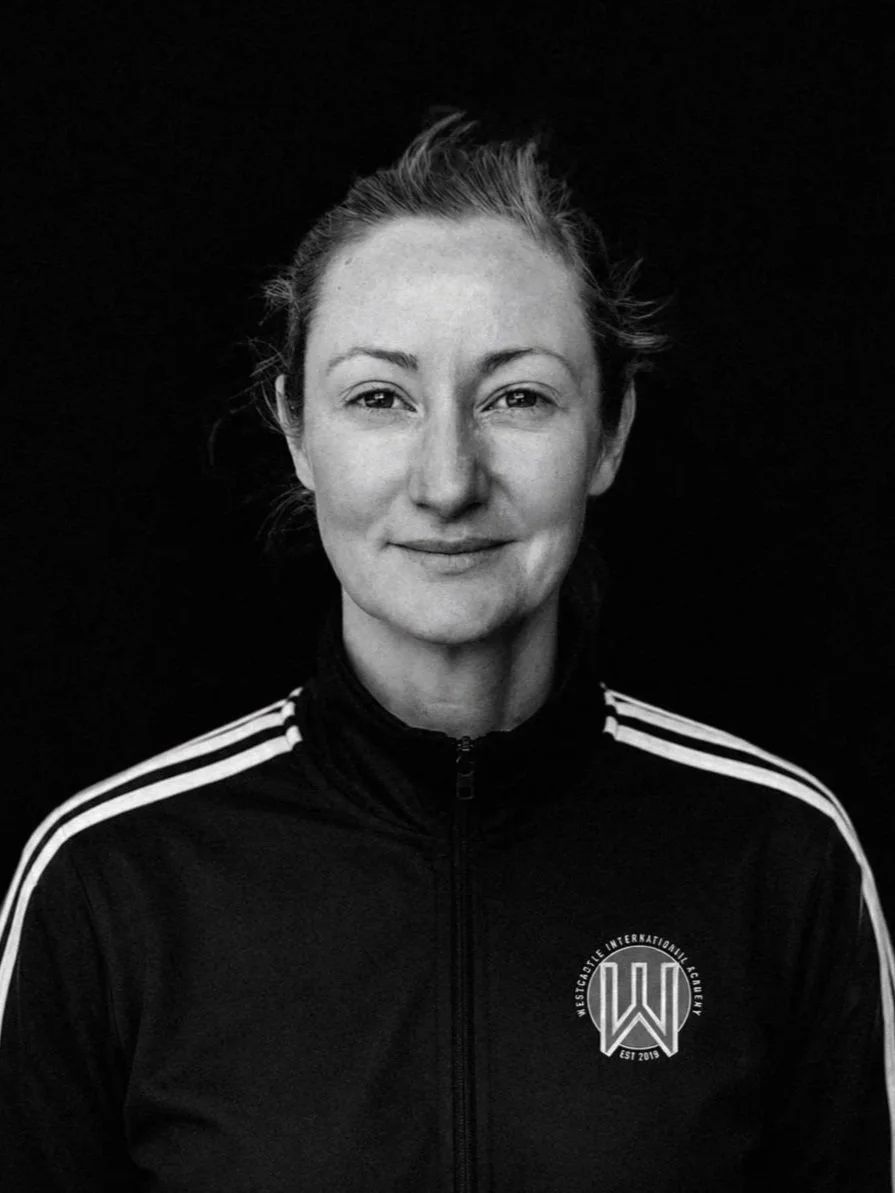 Black and white portrait of a woman with light hair tied back, wearing a black tracksuit jacket with white stripes on the shoulders and a logo that reads 'Wrestleite International Academy' on the chest.