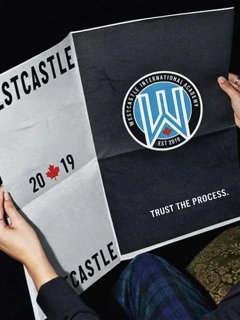 Person holding a black and white poster with the logo of Westcastle International Academy, featuring a blue shield with an 'W' and a red maple leaf, and the text 'Trust the process.'