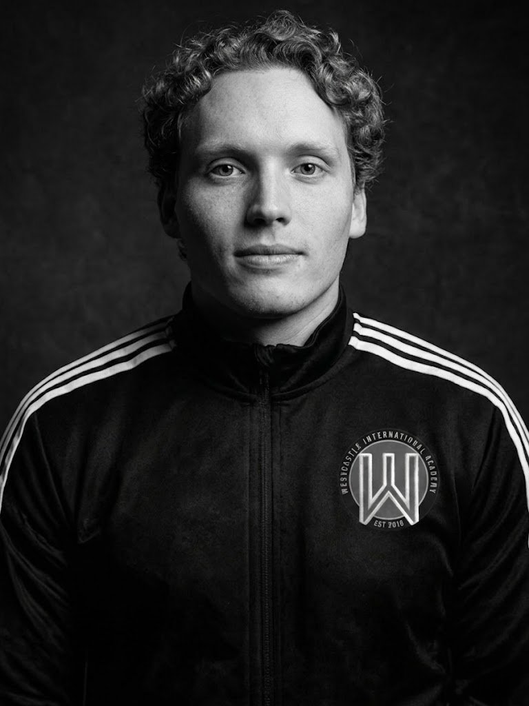 Black and white portrait of a young man with curly hair wearing a sports jacket with a logo on the chest.